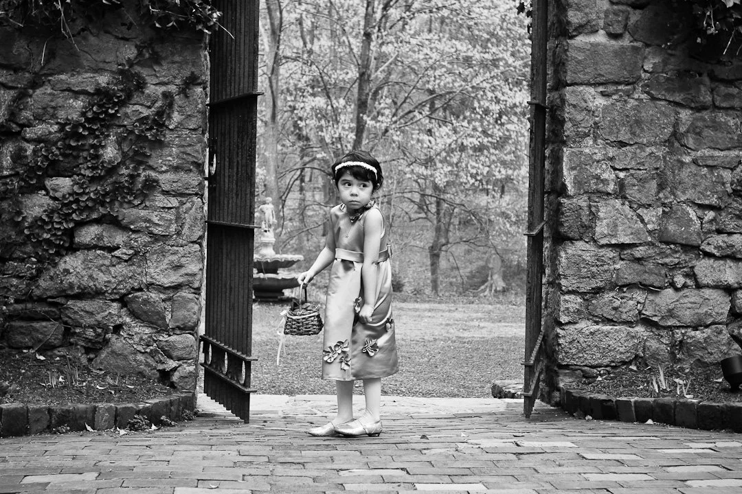 A young girl in a dress holding a small basket stands in an open stone gate, with trees and a fountain visible in the background.