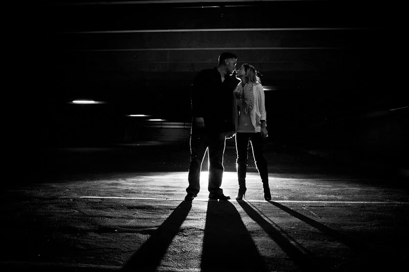 A couple standing close together in the dark, holding hands, with strong backlighting creating long shadows on the ground.
