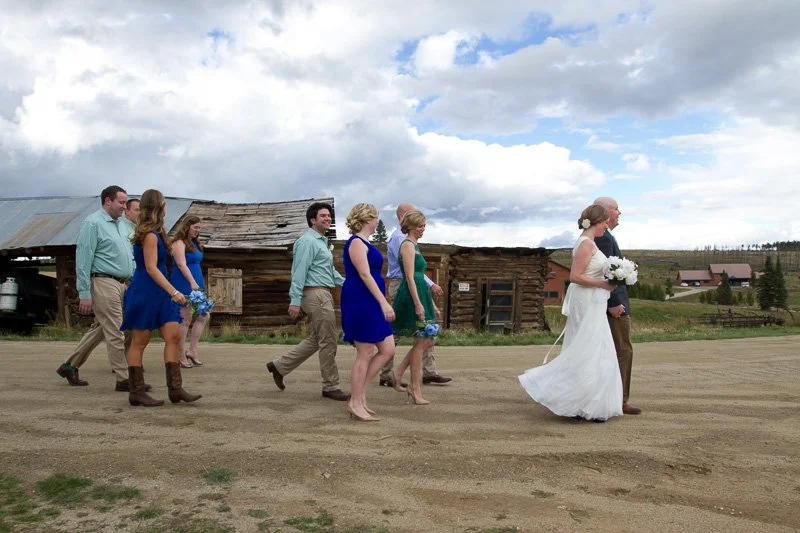 A group of people walking outdoors in a rural setting, with a bride in a white wedding dress leading the way, followed by groom and guests dressed in blue and earth-tone outfits, against a backdrop of rustic buildings and a cloudy sky.
