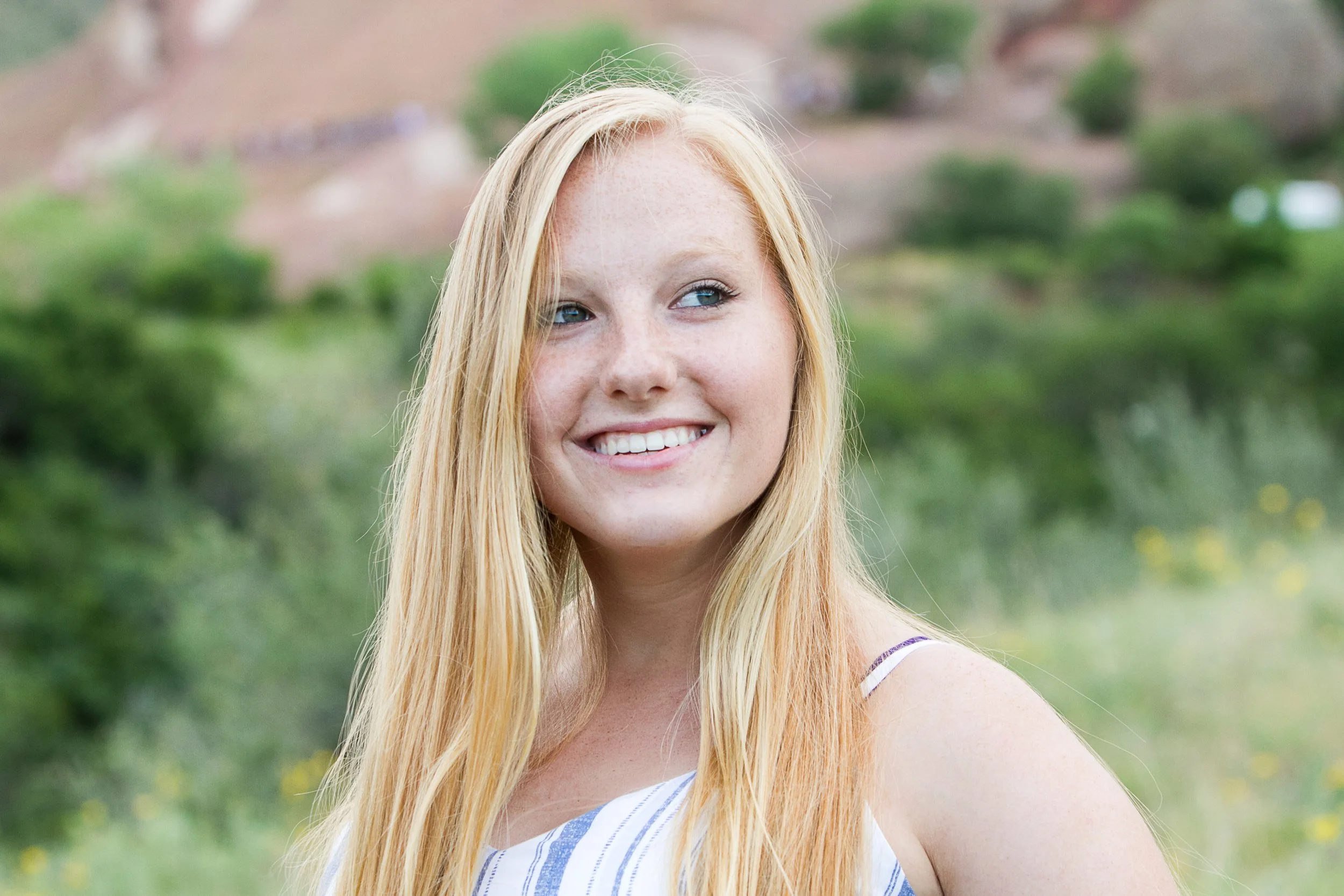 Young woman with long blonde hair smiling outdoors with green trees and hills in the background.