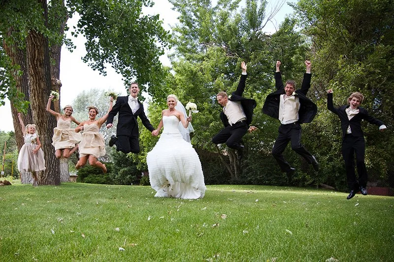 A wedding group photo shows the bride and groom holding hands, surrounded by bridesmaids and groomsmen, all jumping in the air on a grassy lawn with trees in the background.
