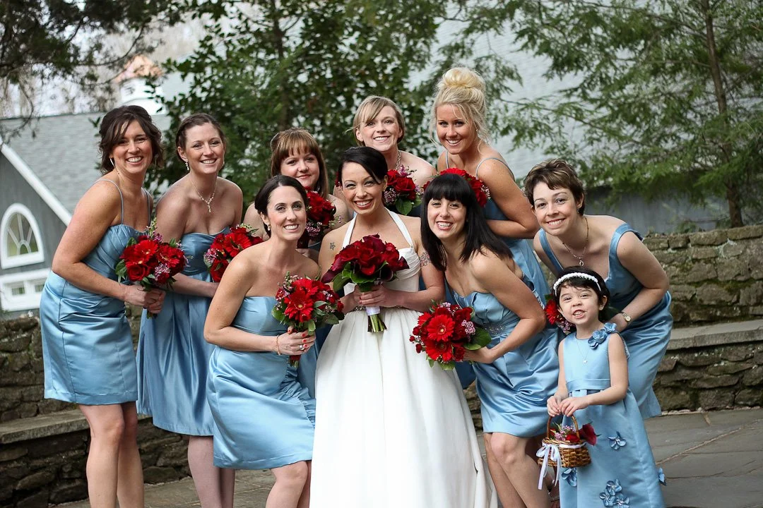 A group of women, including a bride and her bridesmaids, posing outdoors for a wedding photo, holding red bouquets, with trees and a stone wall in the background.