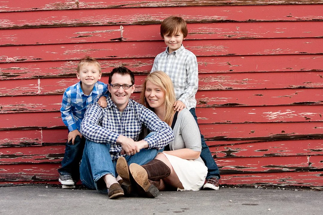 A family of four outdoors leaning against a weathered red wooden wall. The father, mother, and two young boys are smiling at the camera.