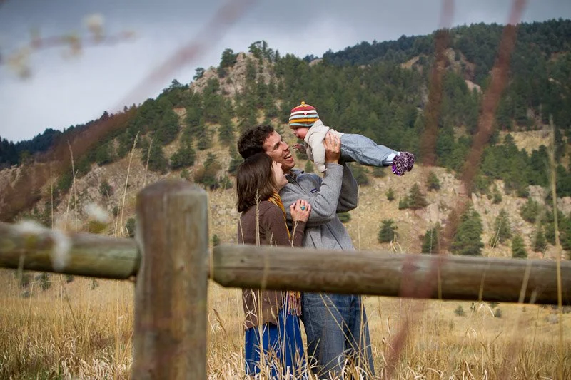 Family outdoors in a grassy field with hills in the background, father lifting a baby wearing a hat, mother looking at the baby.