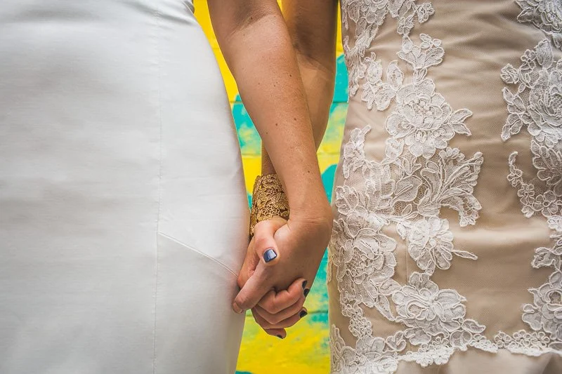 Two women holding hands, one wearing a white dress and the other in a beige lace dress, against a colorful background.