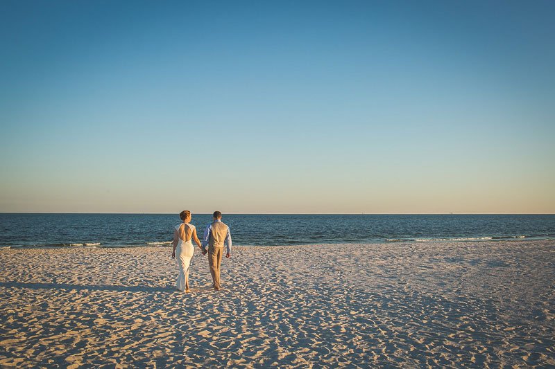 A couple dressed in white and beige walking hand in hand along the beach at sunset.
