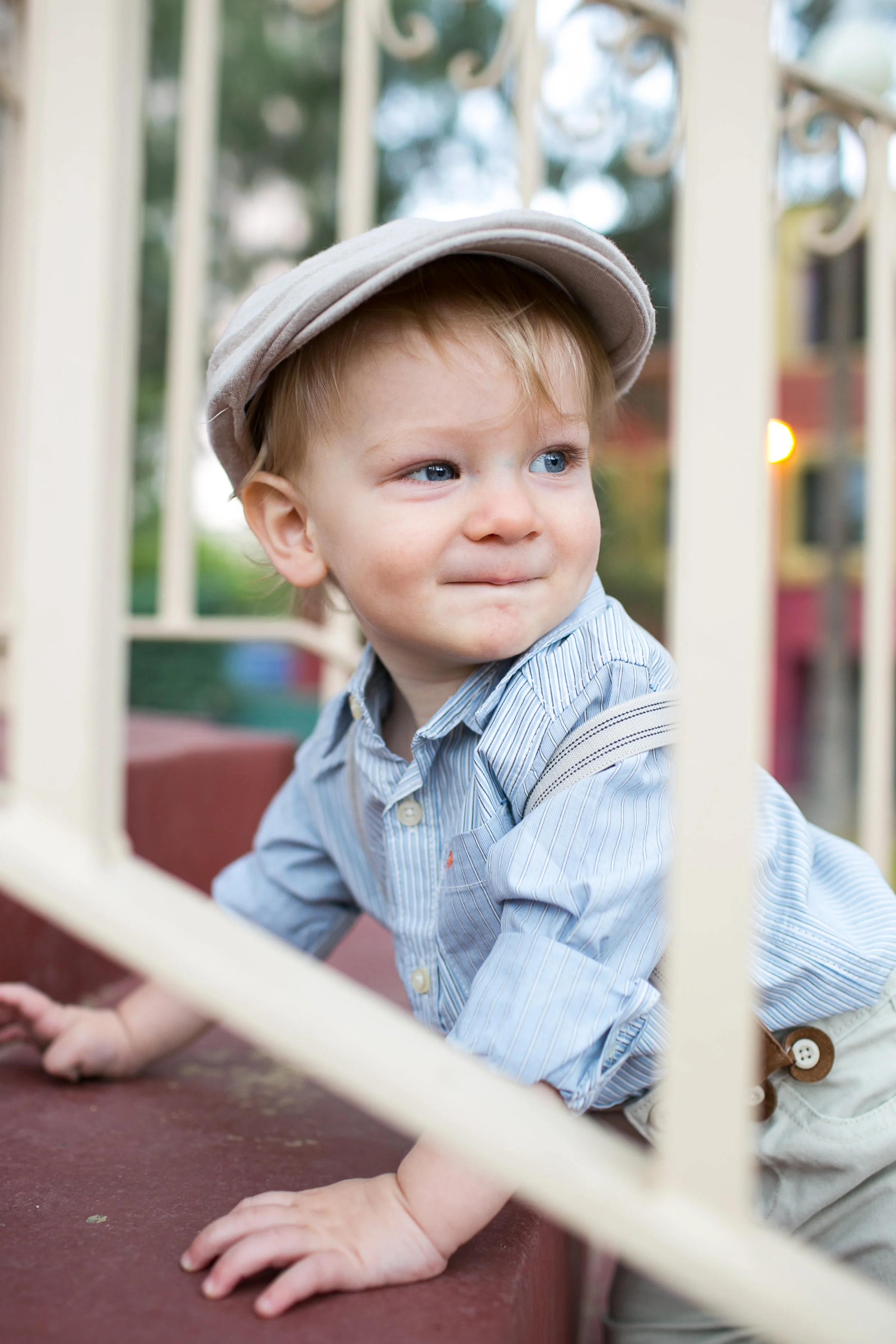 Young boy wearing a beige hat and a light blue striped shirt, crawling on a red surface with spatial metal bars in the foreground, outdoors in a park or playground.