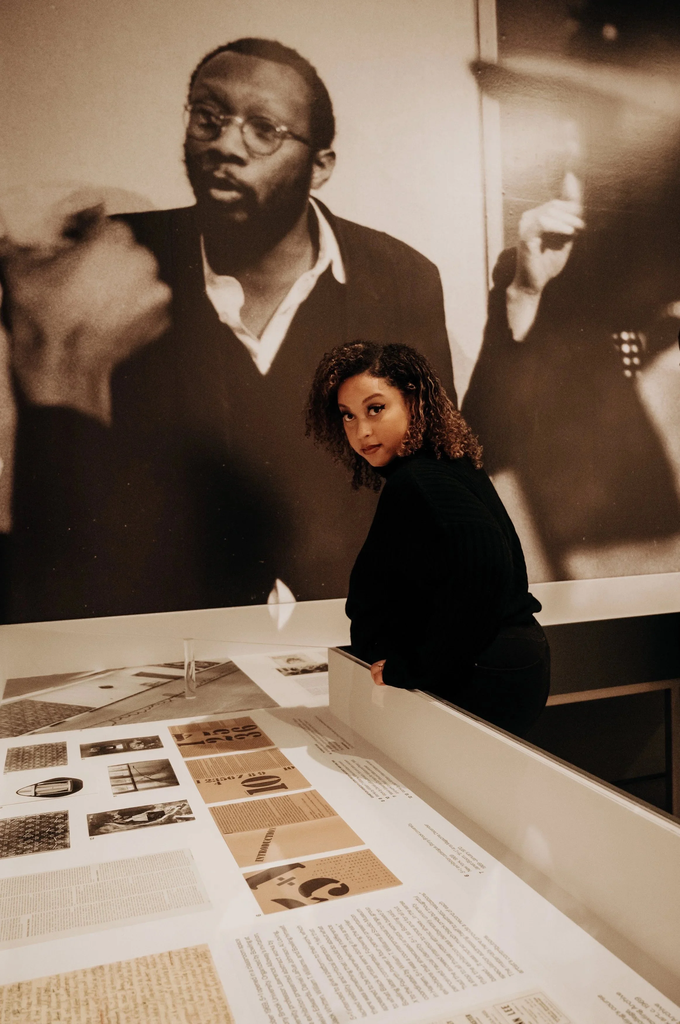 A woman with curly hair and a black outfit standing and looking at historical documents and photographs on display, with a large black-and-white photograph of a man with glasses and a beard in the background.