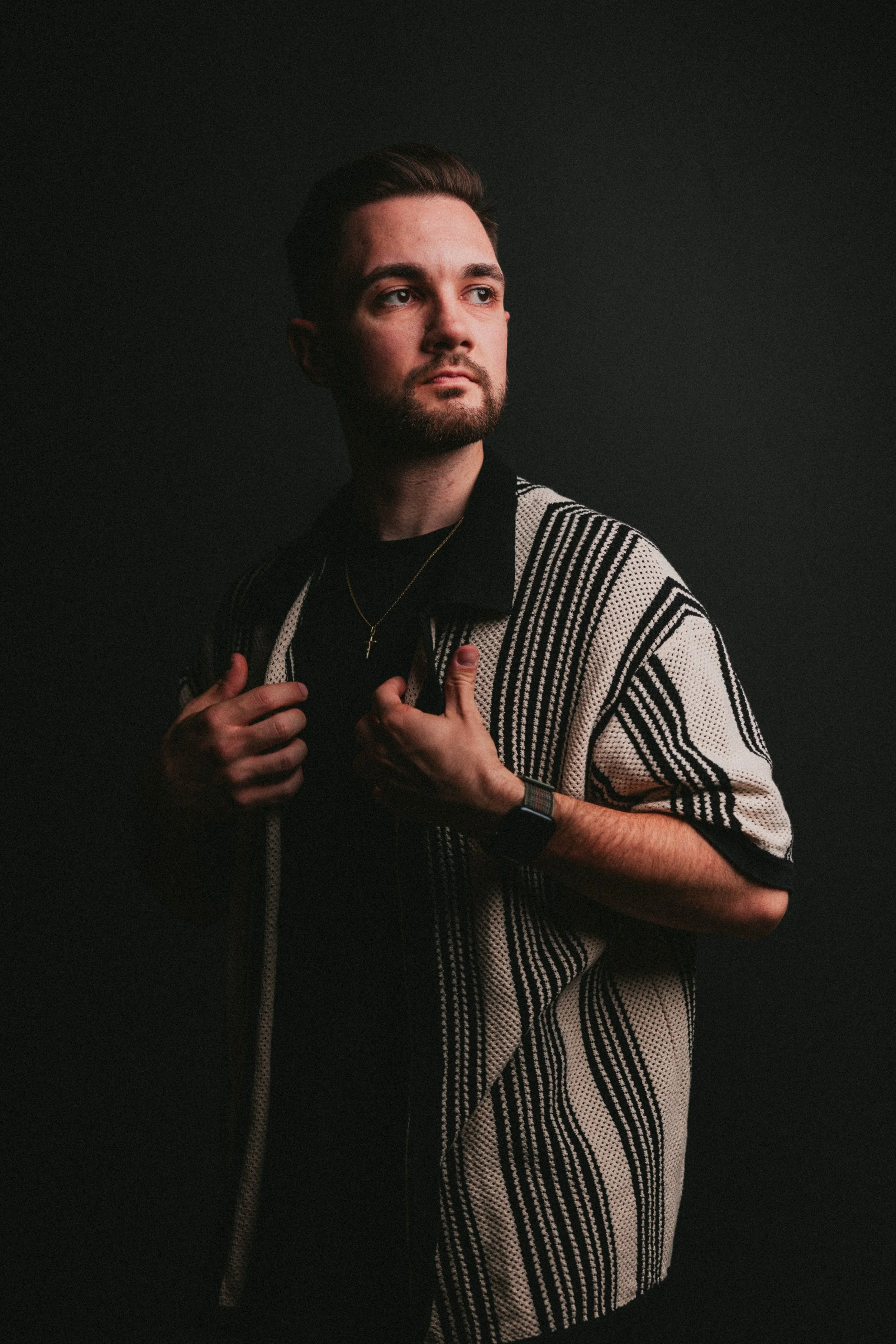 Ein junger Mann mit dunklem Haar, Bart und dunklem Blick, trägt eine schwarze Kleidung und einen mantel mit schwarzen und weißen Streifen vor einem dunklen Hintergrund. Er schaut nach rechts und hält den Mantel mit beiden Händen.