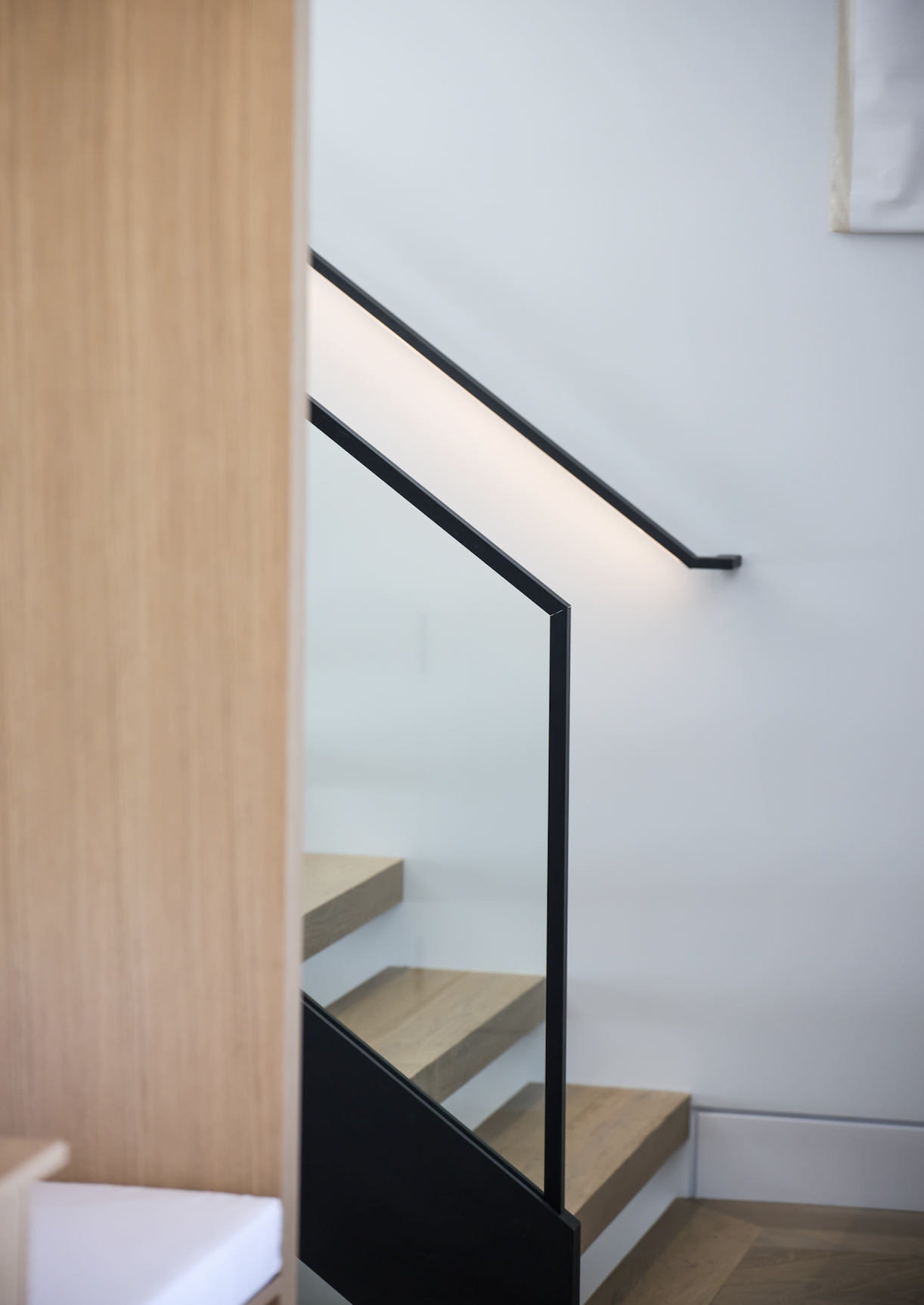Interior view of a staircase with wooden stairs, a black metal handrail, and a white wall. Part of a wooden wall or furniture is visible on the left side.