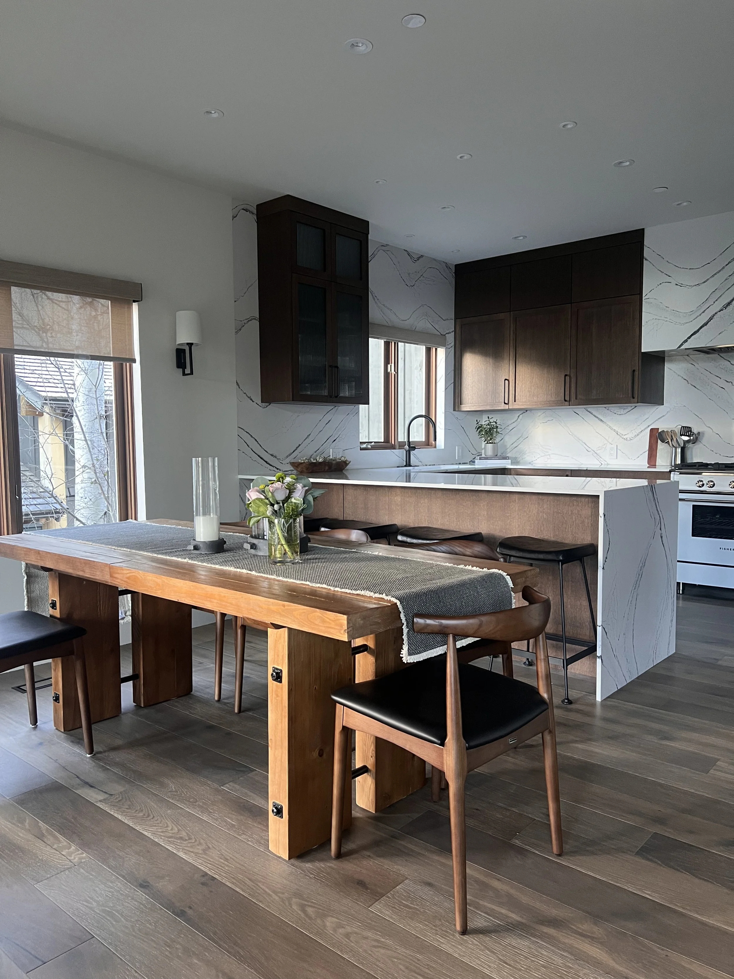 Modern kitchen with wooden dining table, black chairs, marble backsplash, and a window with a view of outdoor trees