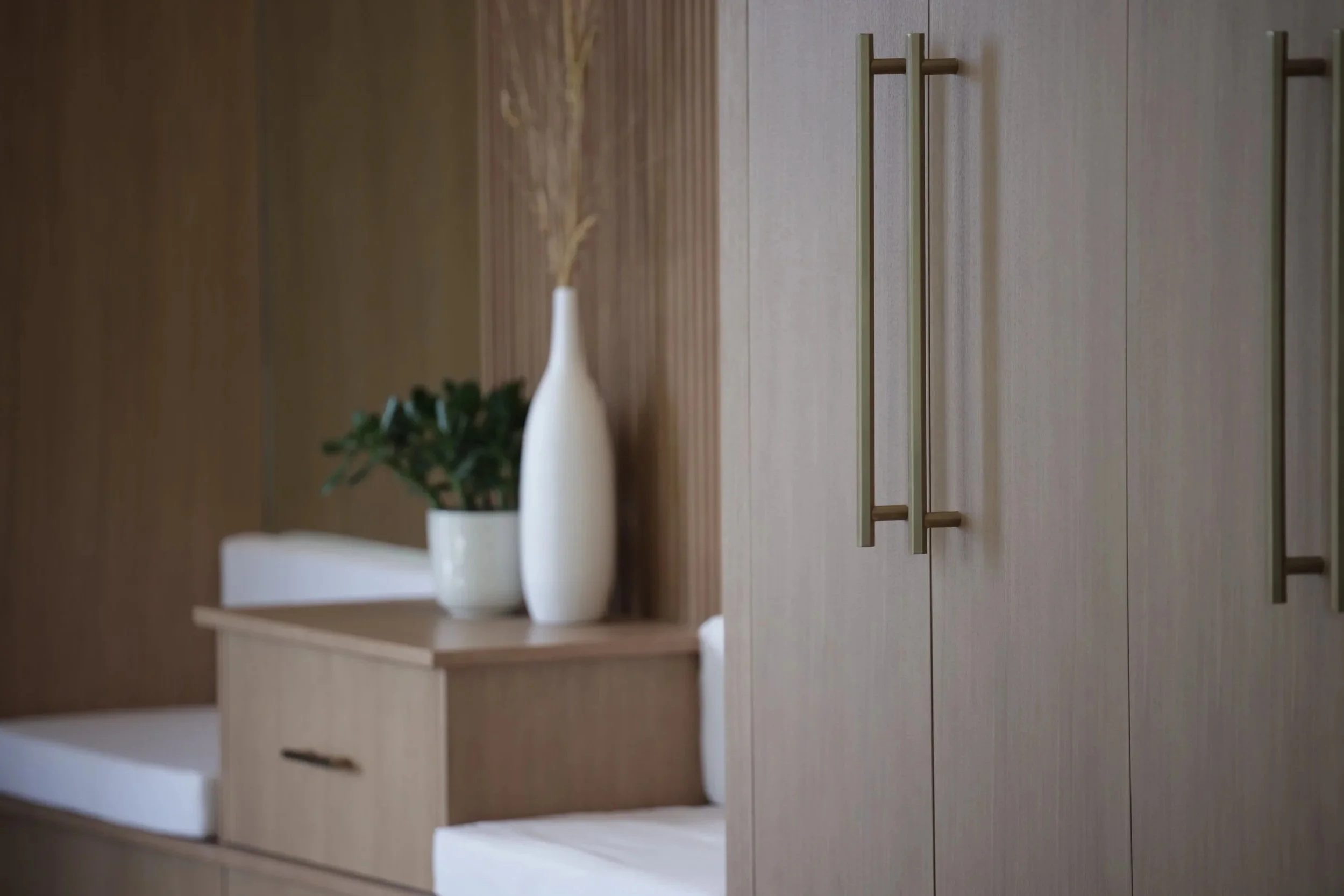 Close-up of wooden cabinet doors with brass handles in a modern interior, with a table holding a white vase with dried floral arrangement and a small potted plant visible in the background.