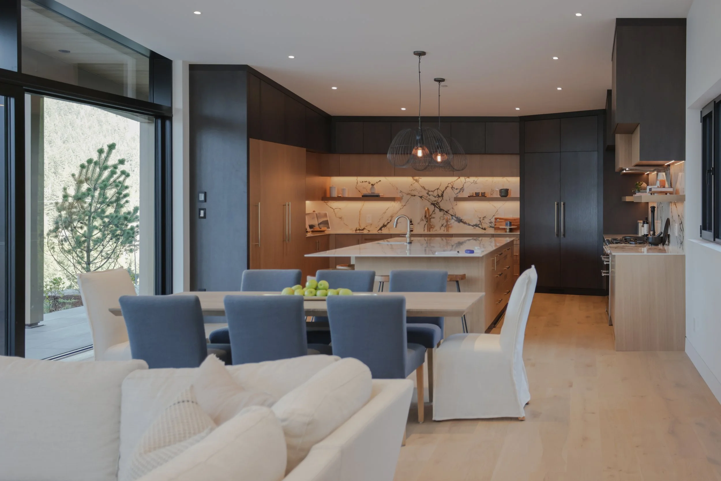 Modern open-concept kitchen and dining area with a large marble-topped island, dark and light wood cabinetry, a dining table with gray and white chairs, and sliding glass doors revealing a tree outside.