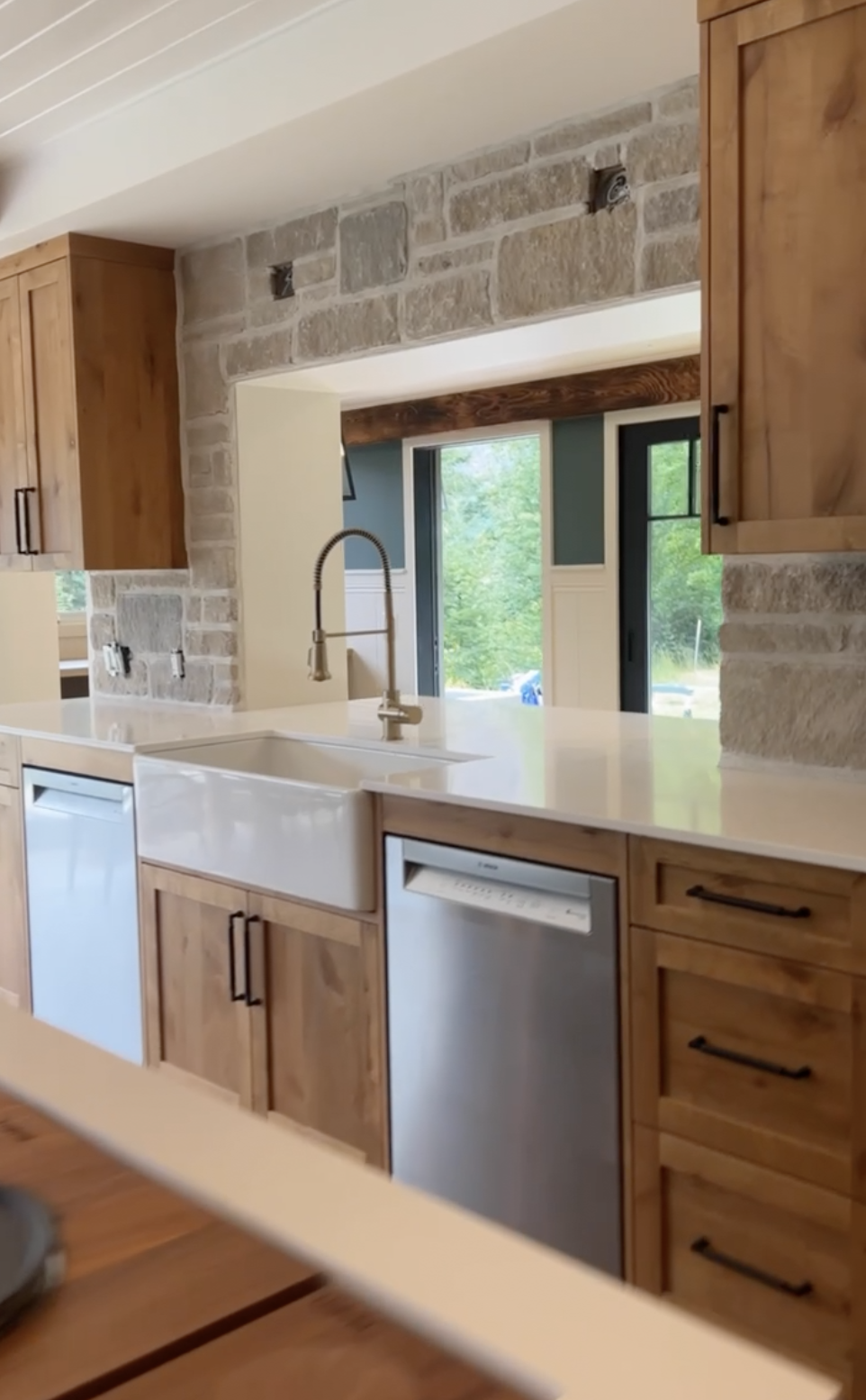Kitchen with wooden cabinets, white countertop, farmhouse sink, stainless steel dishwasher, stone wall, large window, and outdoor view.