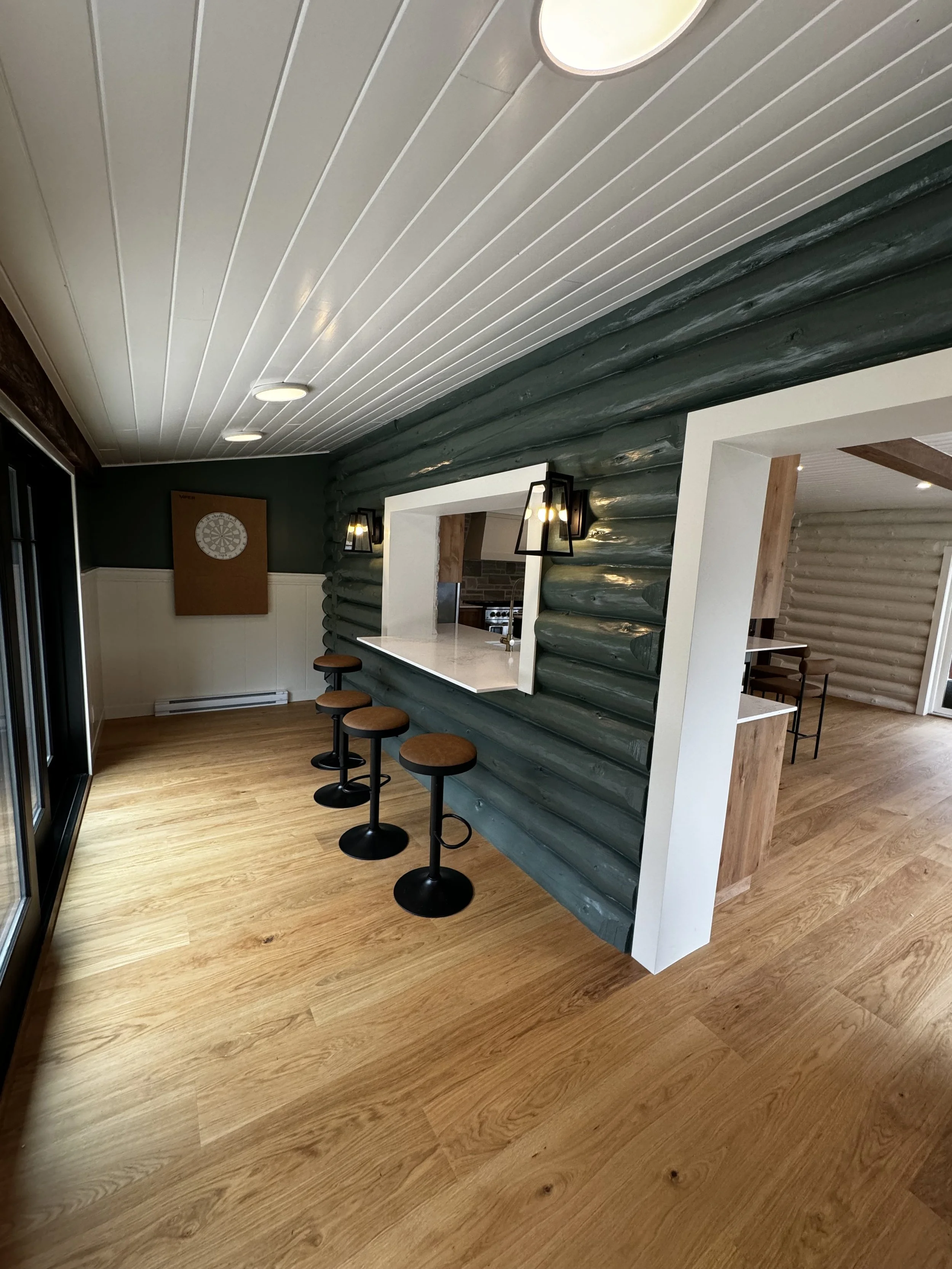 Interior of a room with wooden floors, green log wall, and a counter opening to the kitchen. Four bar stools are placed along the counter. Black wall-mounted lanterns are on either side of the window opening.