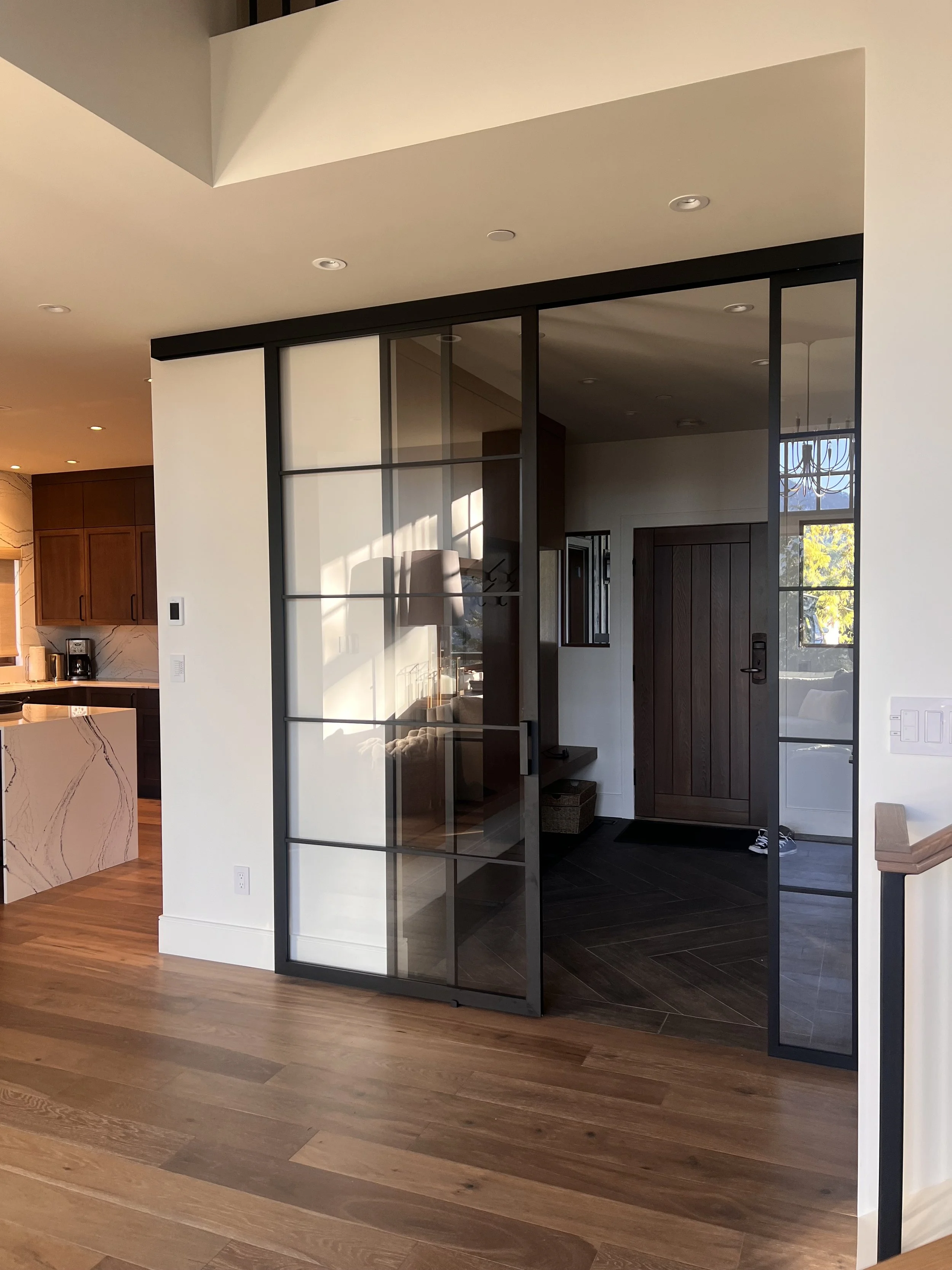 The image shows an interior view of a modern home entryway with a black-framed glass partition door. The door is partially open, revealing a space with dark wood flooring and a front door. To the left, part of a kitchen with wooden cabinets and an is
