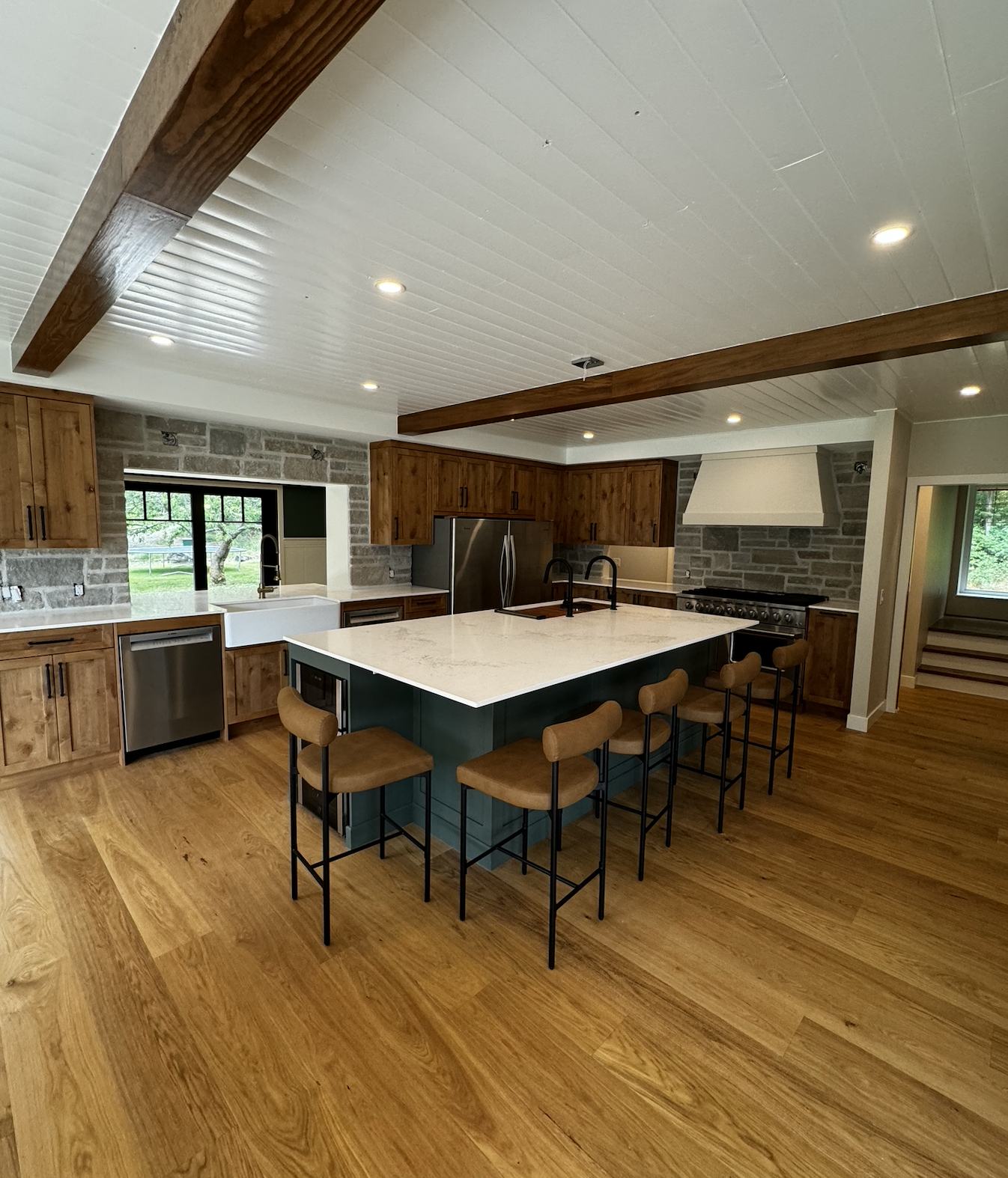 Modern kitchen with wooden cabinets, a large central island with a white countertop, bar stools, stainless steel appliances, a stone accent wall, and wooden flooring.