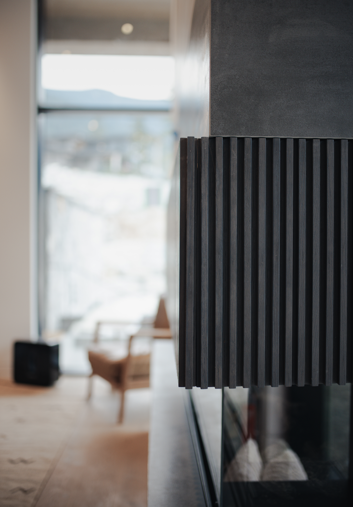 Close-up of a modern black textured wall with a blurred background featuring a beige armchair and a small black speaker near a large window.