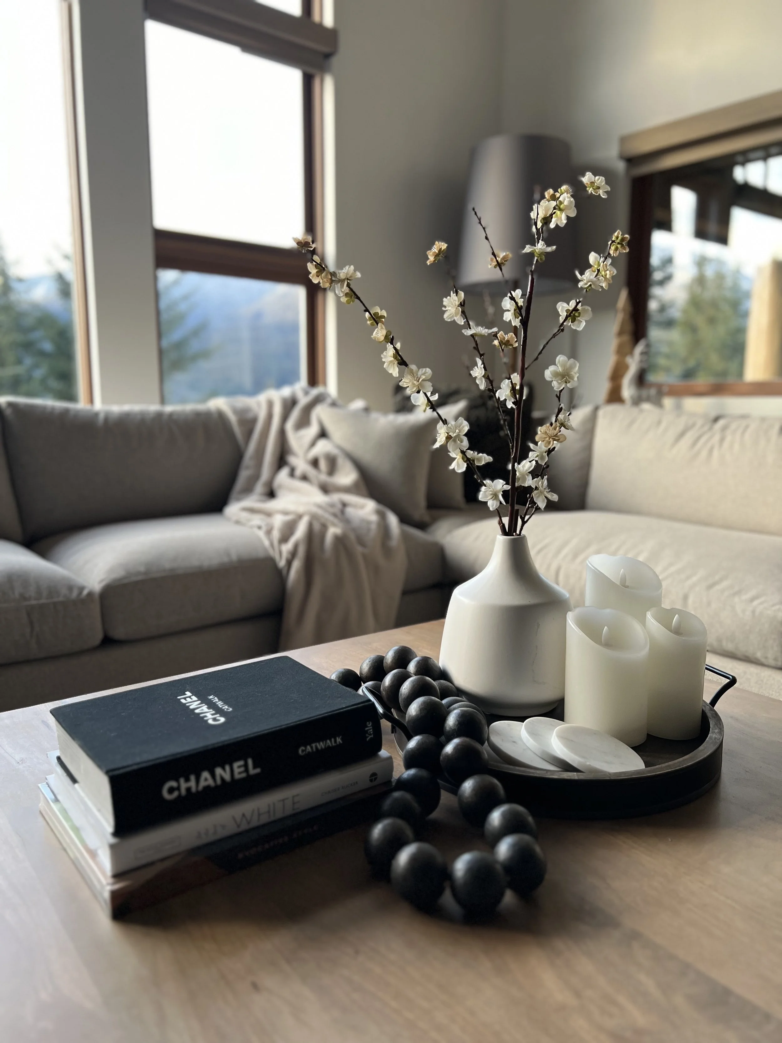 A cozy living room with a wooden table featuring a white vase with branches of white blossoms, three white candles, black beads, and a stack of fashion and lifestyle books. In the background, there is a beige sofa with a blanket and a window showing 
