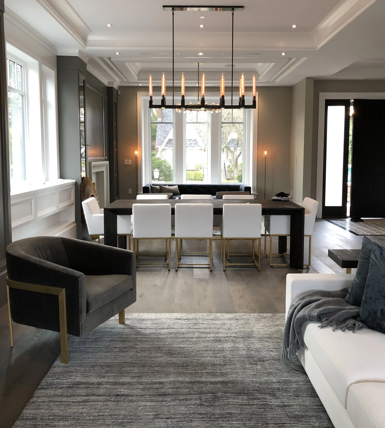 A modern dining and living room area with large windows, a black dining table with white chairs, dark gray walls, a white sofa with pillows, a gray armchair, and a large rectangular chandelier hanging from the ceiling.