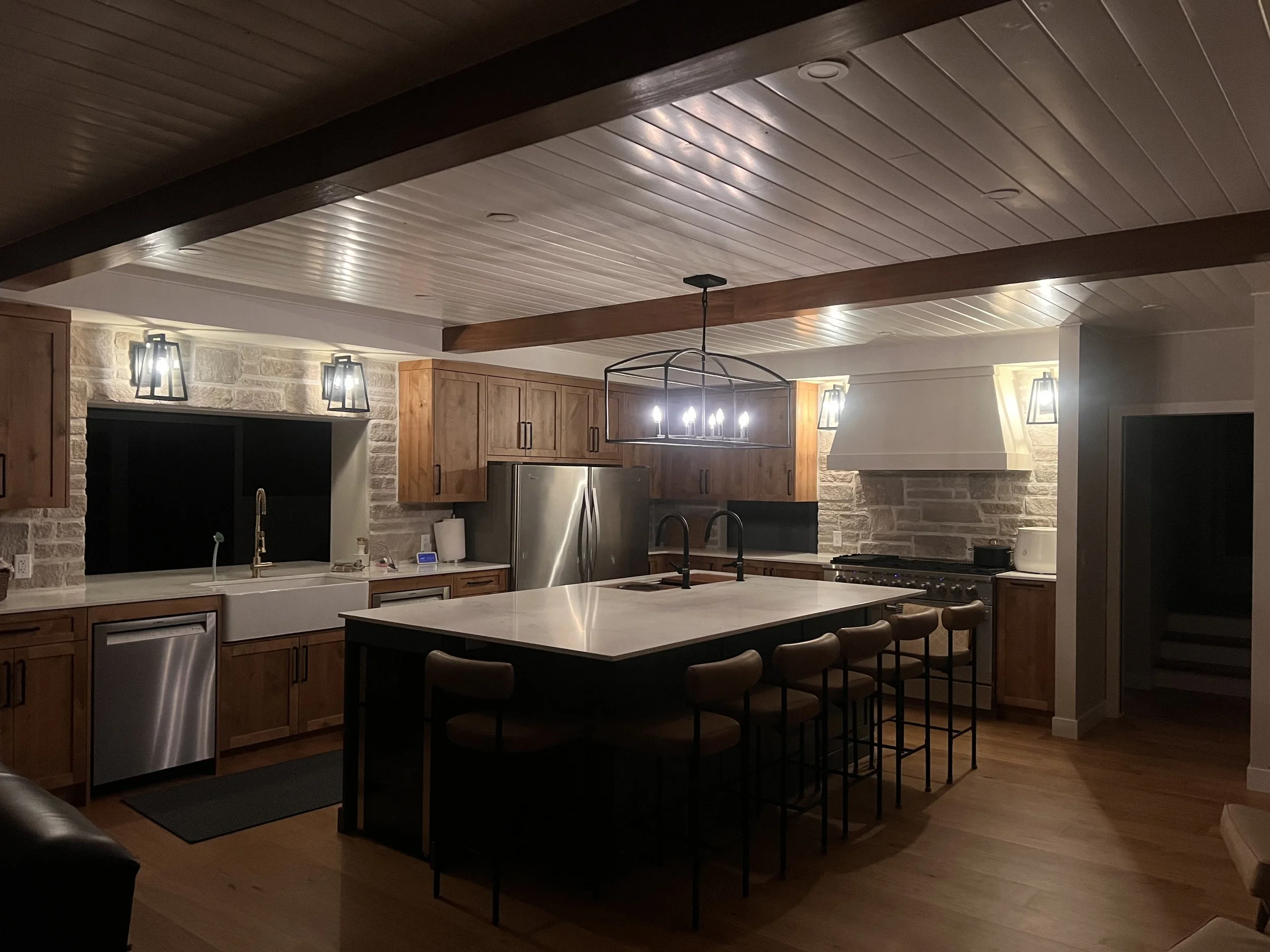 Modern kitchen with stone accent walls, wooden cabinets, stainless steel appliances, and a large island with bar stools, illuminated by wall-mounted and ceiling lighting.