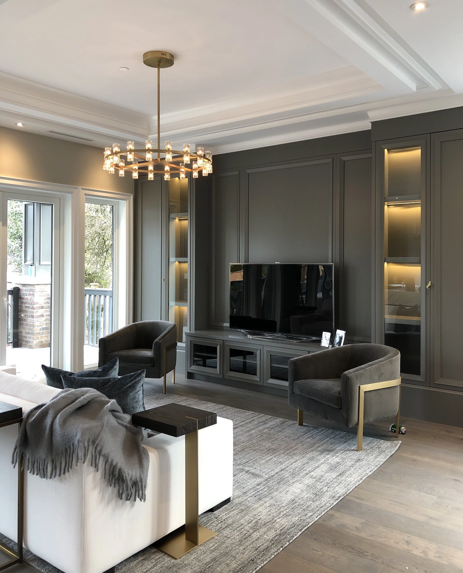 Modern living room with a gray wall unit, a flat-screen TV, two gray armchairs, a white sofa with gray and black pillows, a gray blanket, a chandelier, and sliding glass doors leading to an outdoor balcony.