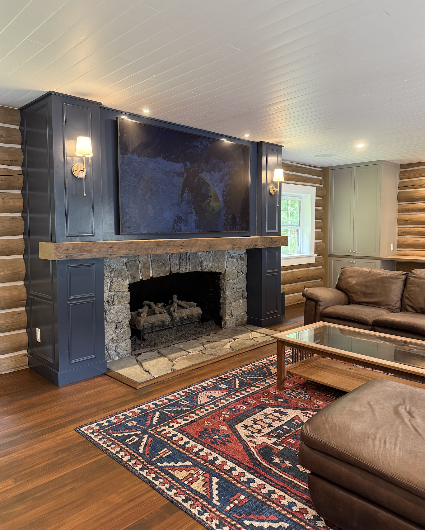 Living room with log cabin walls, a stone fireplace with a wooden mantle, a large mounted flat-screen TV showing a person kayaking, a leather sofa, a glass-top coffee table, a colorful patterned rug, and a window with natural light.