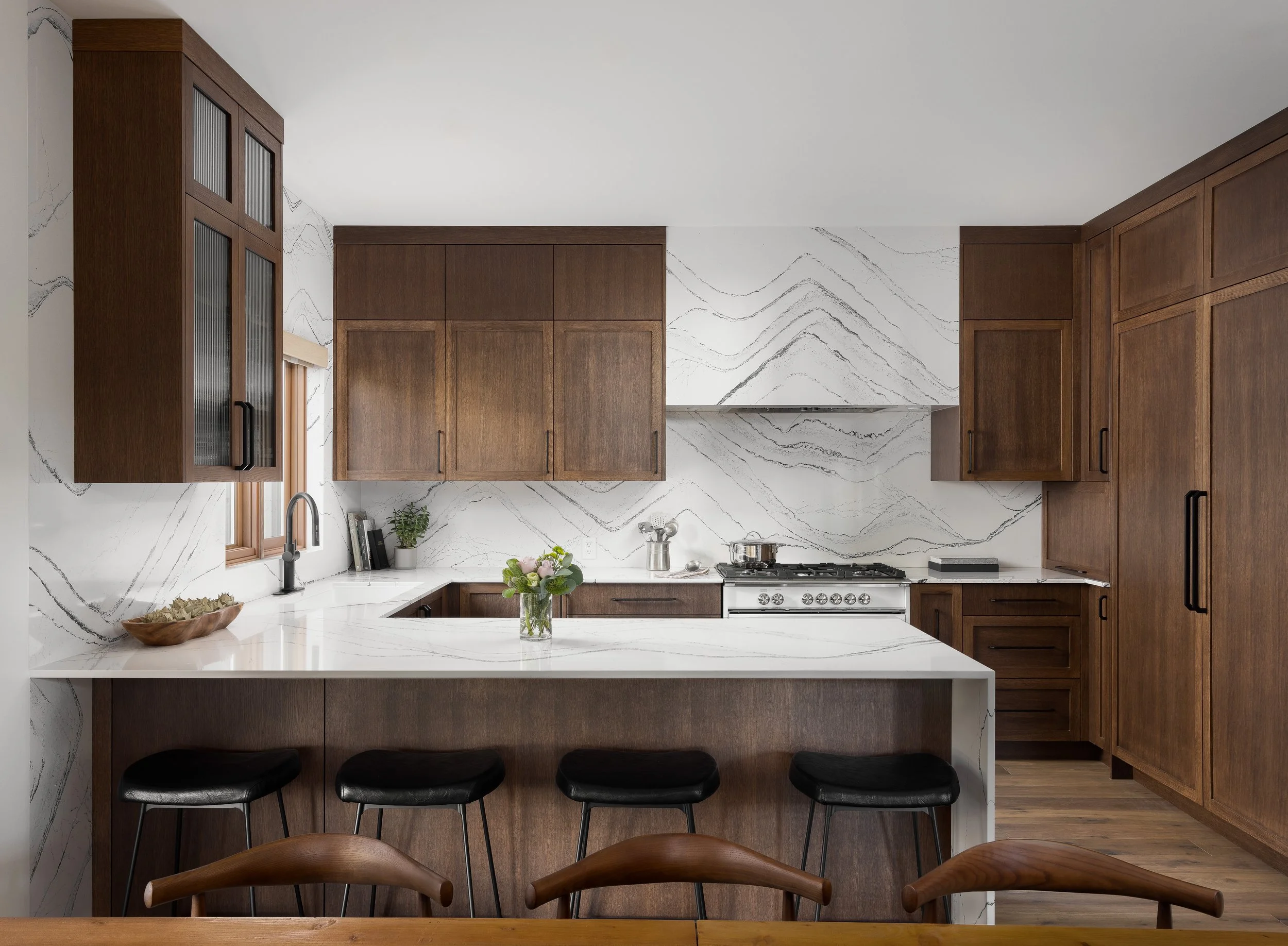 Modern kitchen with white marble countertops and backsplash, wooden cabinets, black barstools, and a window above the sink.