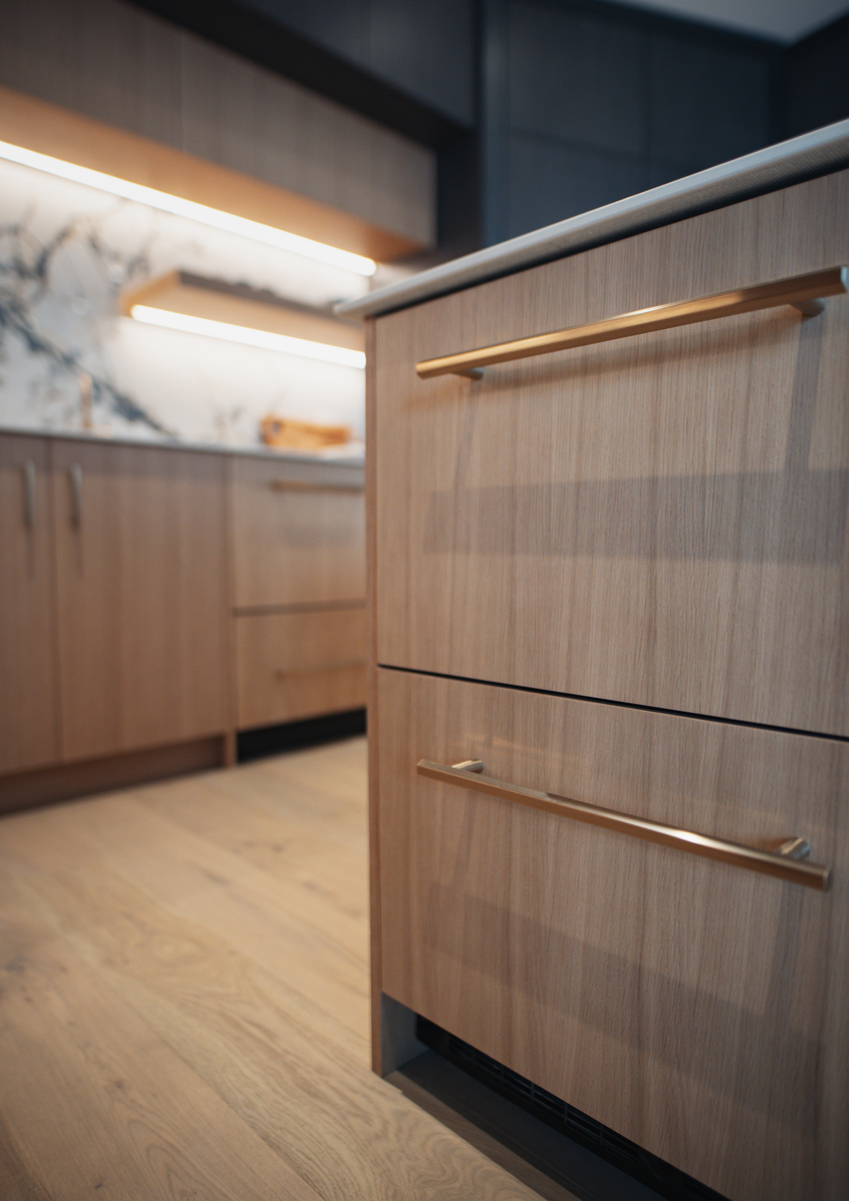 Close-up of a wooden kitchen cabinet with gold handles, showing part of the kitchen's modern interior with a marble backsplash and wooden flooring.