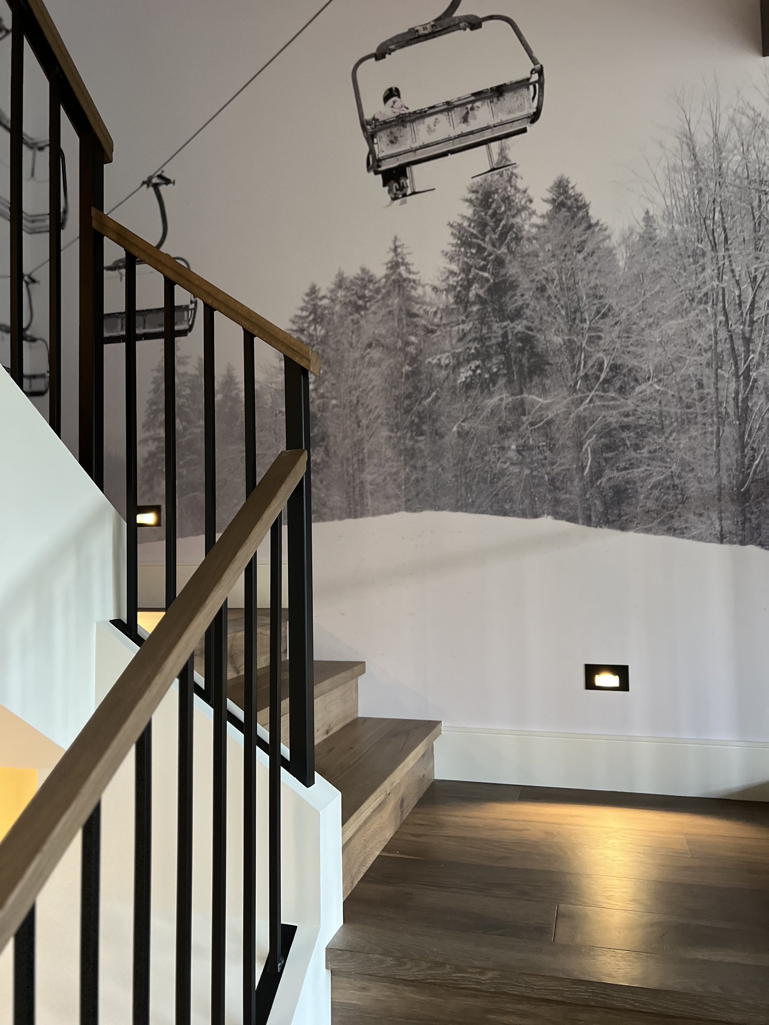 Interior scene of a staircase with wooden steps and black railing, overlooking a wall mural of a snowy forest with ski lift chairs in the background.