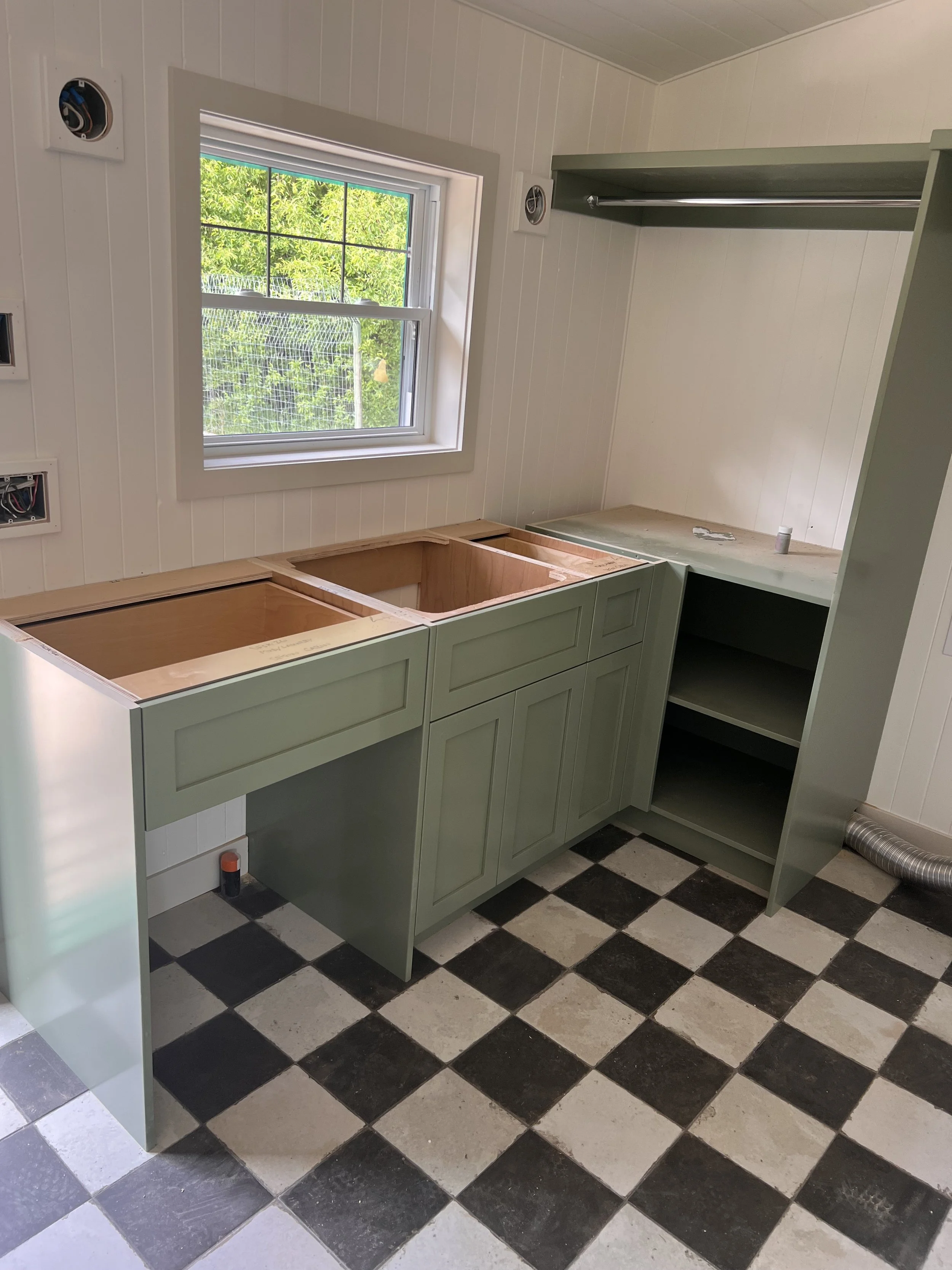 A kitchen under renovation with light green cabinetry, a window with white trim, and black and white checkered floor tiles.