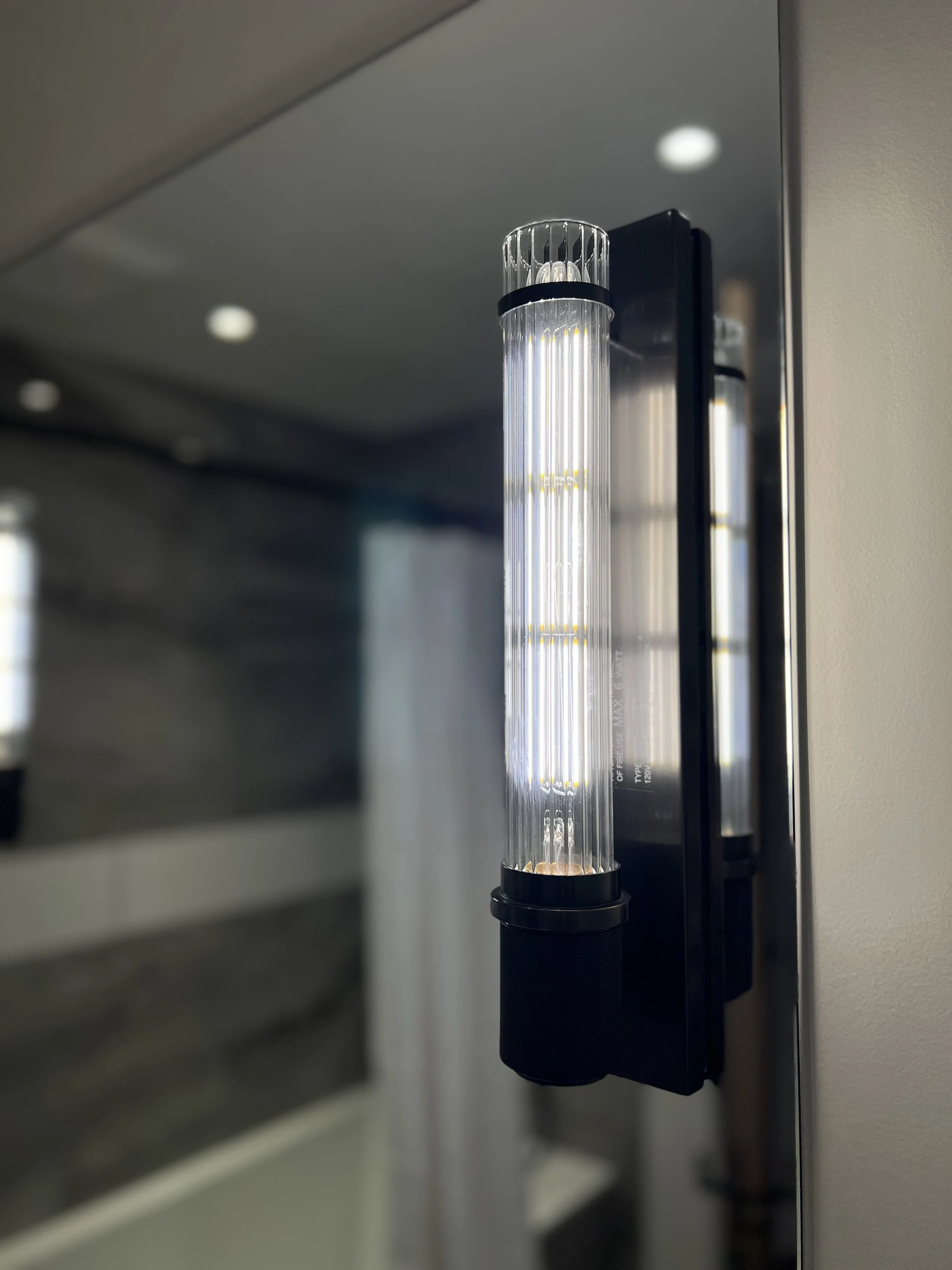Close-up of a modern, vertical light fixture mounted on a wall, with a reflective surface and a blurred background.