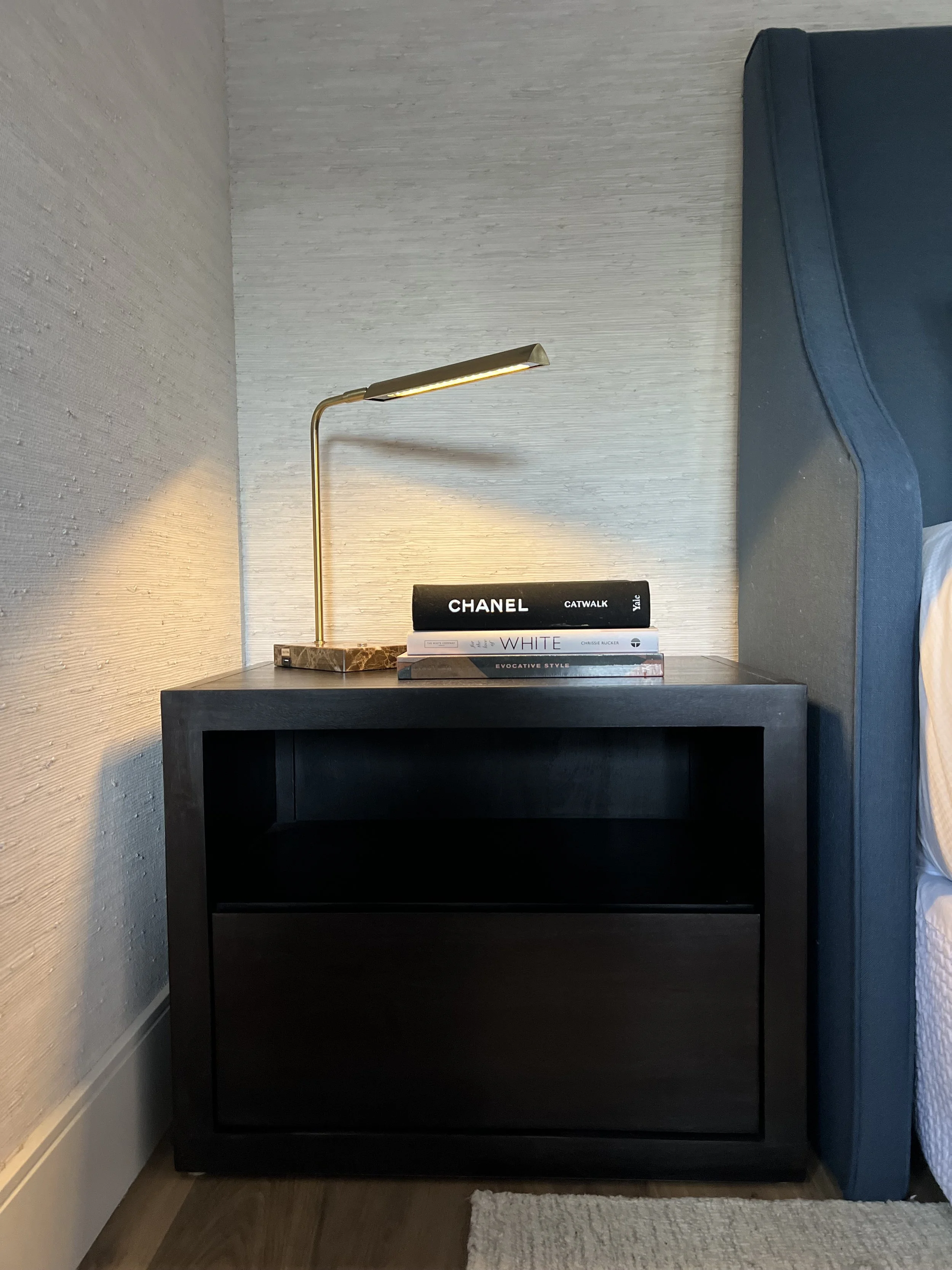 Black nightstand with a gold desk lamp and a stack of three books, placed beside a blue upholstered bed with white bedding, in a hotel room.