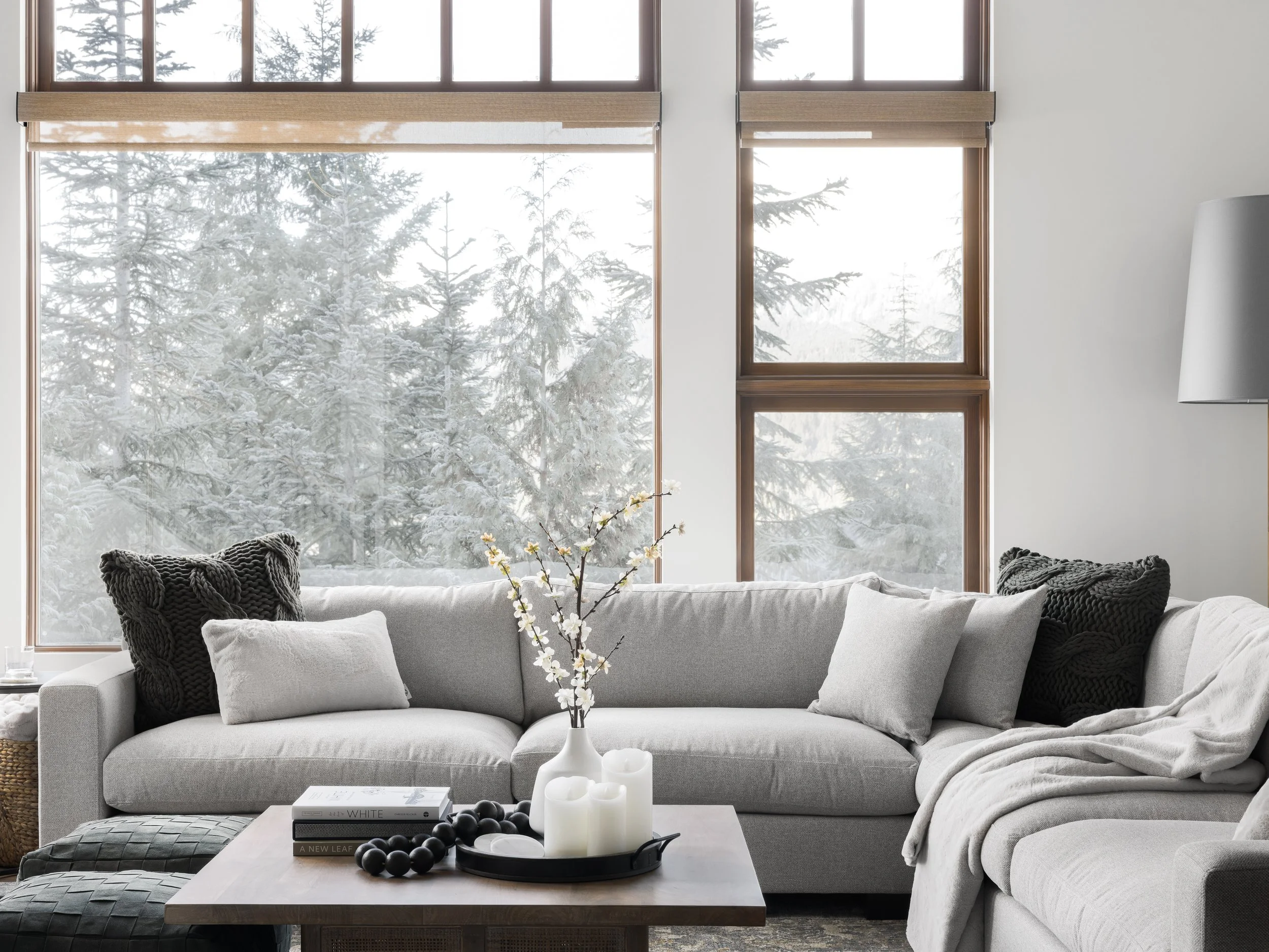 Living room with a large sofa, decorative pillows, a coffee table with candles, flowers, and books, and large windows showing snowy trees outside.