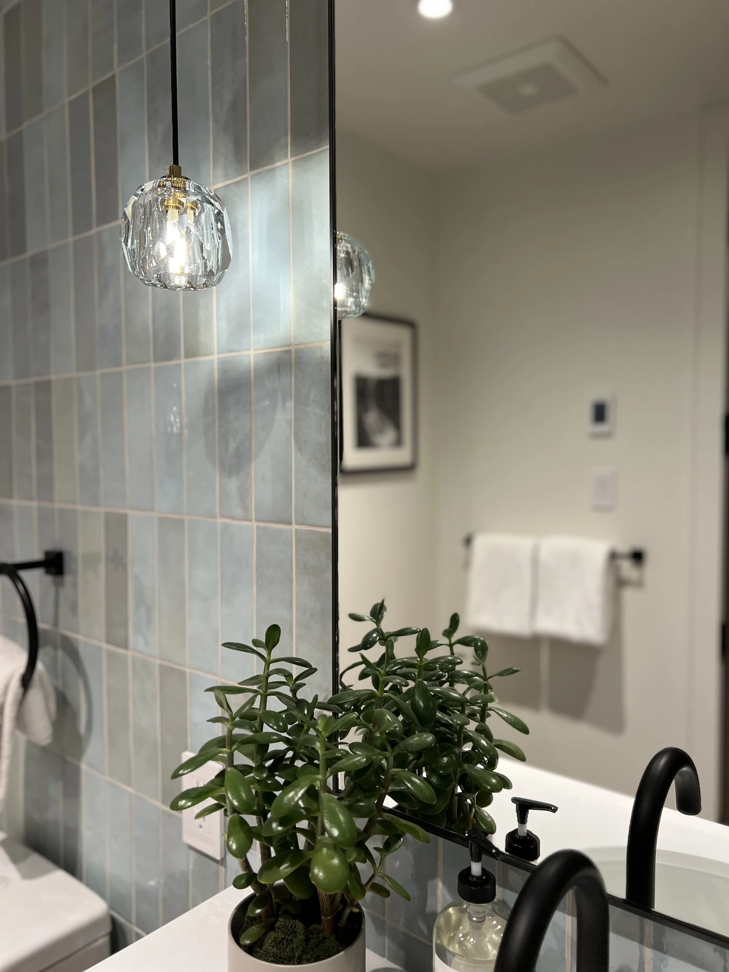 Inside a bathroom with a mirror, a hanging light fixture, a green potted plant on the counter, a soap dispenser, a black faucet, a towel rack with white towels, and framed art on the wall.