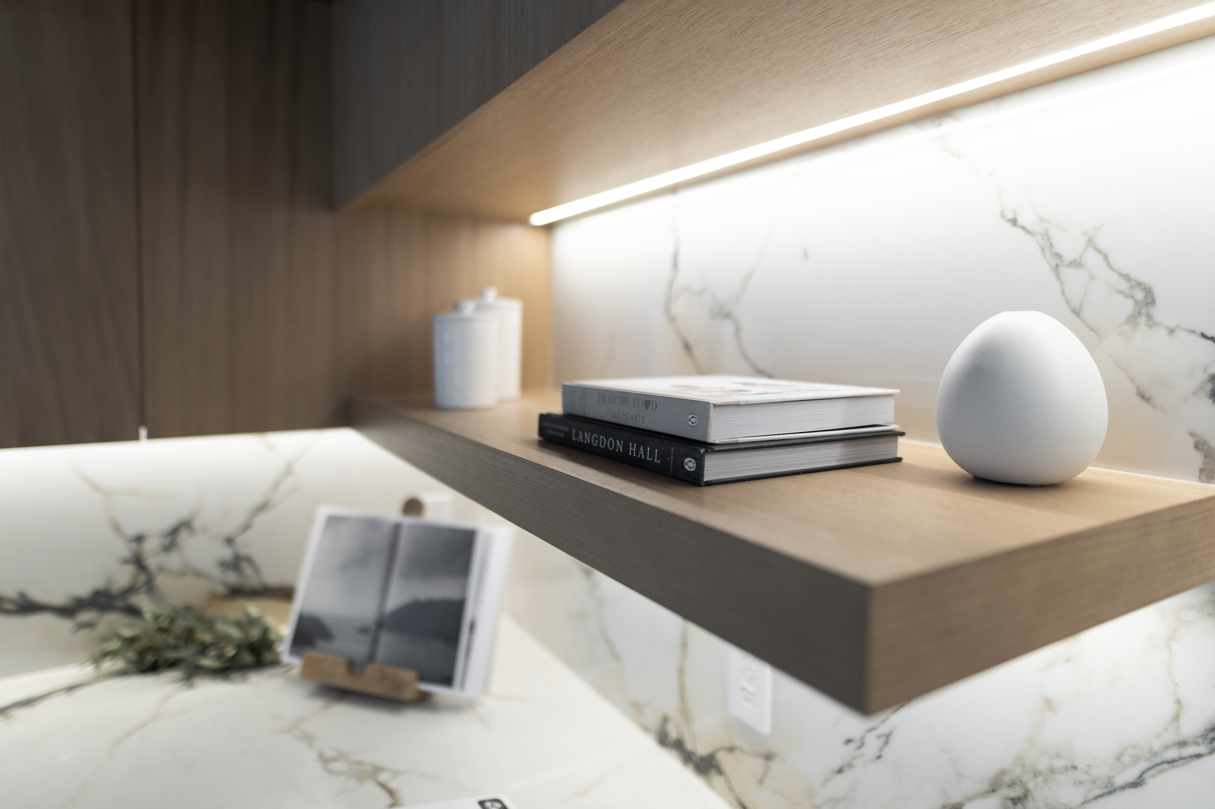 Close-up of a wooden shelf with a white decorative object and stacked cookbooks, with a marble wall behind it.