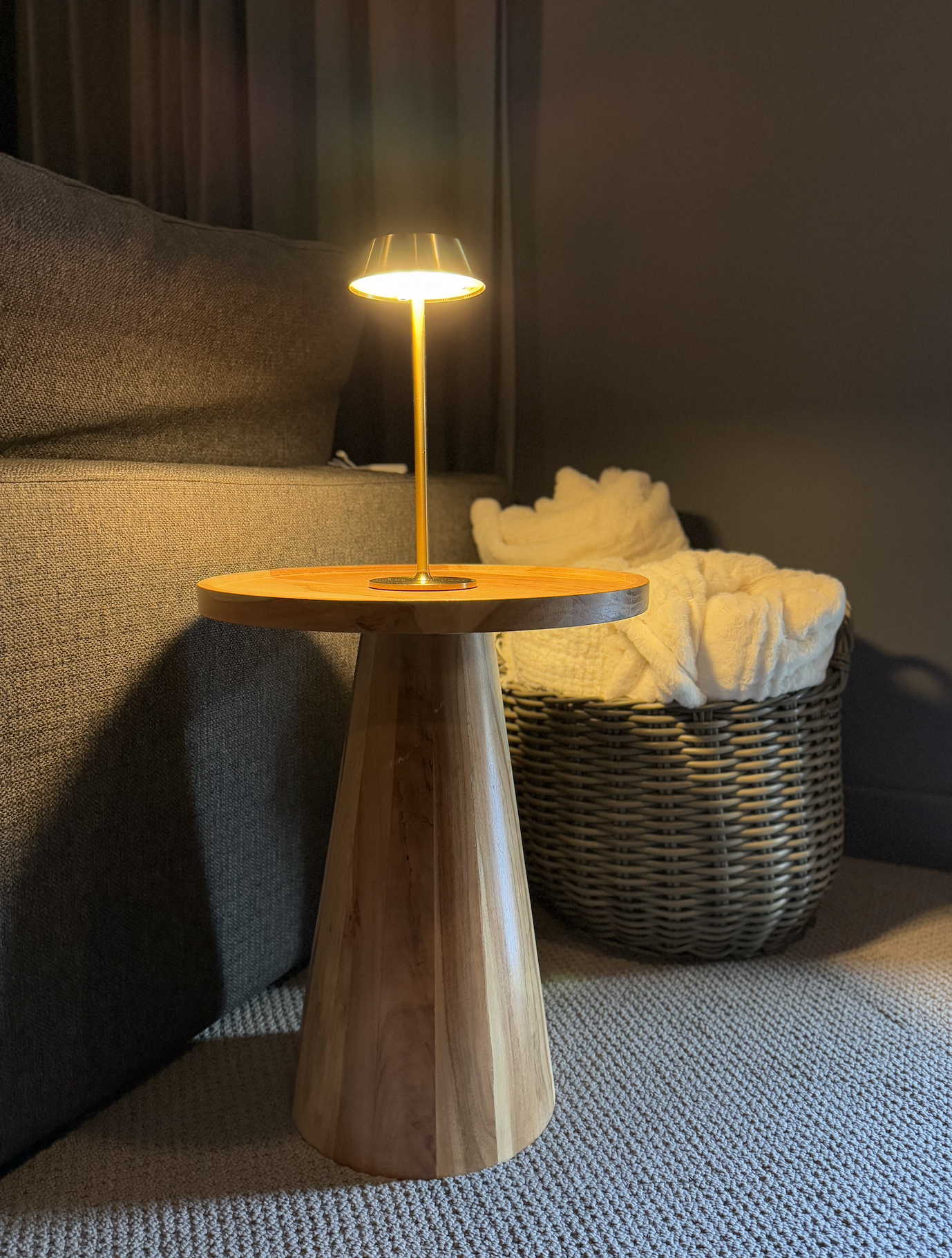 A small wooden side table with a tiny yellow lamp on top, beside a sofa with dark-colored cushions and a laundry basket filled with white towels in a cozy room.
