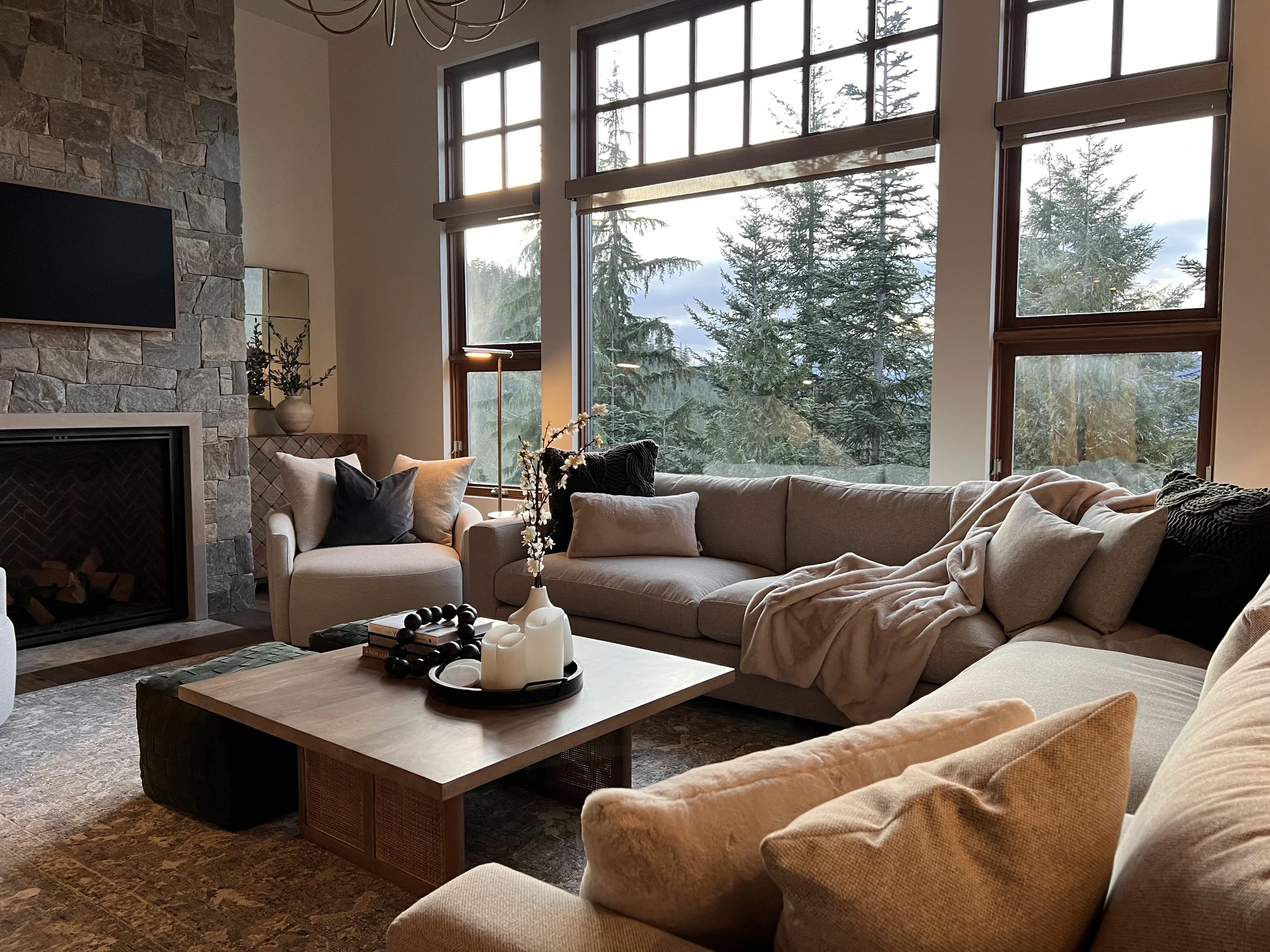 Living room with large windows overlooking trees, beige sectional sofa with pillows and blanket, wood coffee table with candles, armchair, stone fireplace, and wall-mounted television.