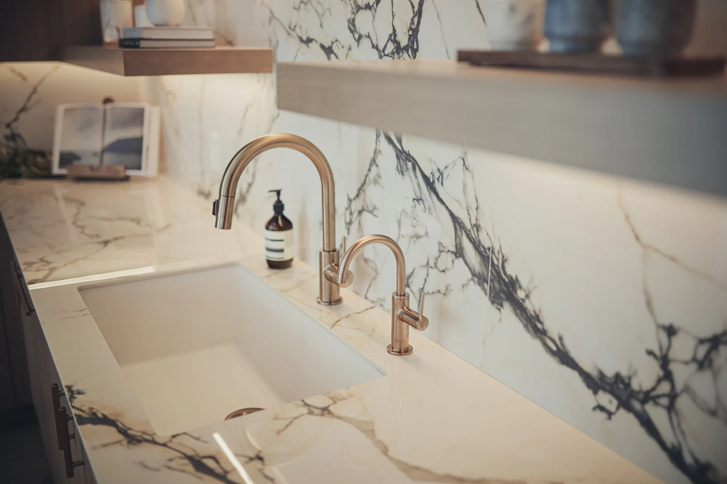 A kitchen sink with a marble countertop and backsplash, featuring two rose gold faucets, a soap dispenser, and framed pictures on the counter.