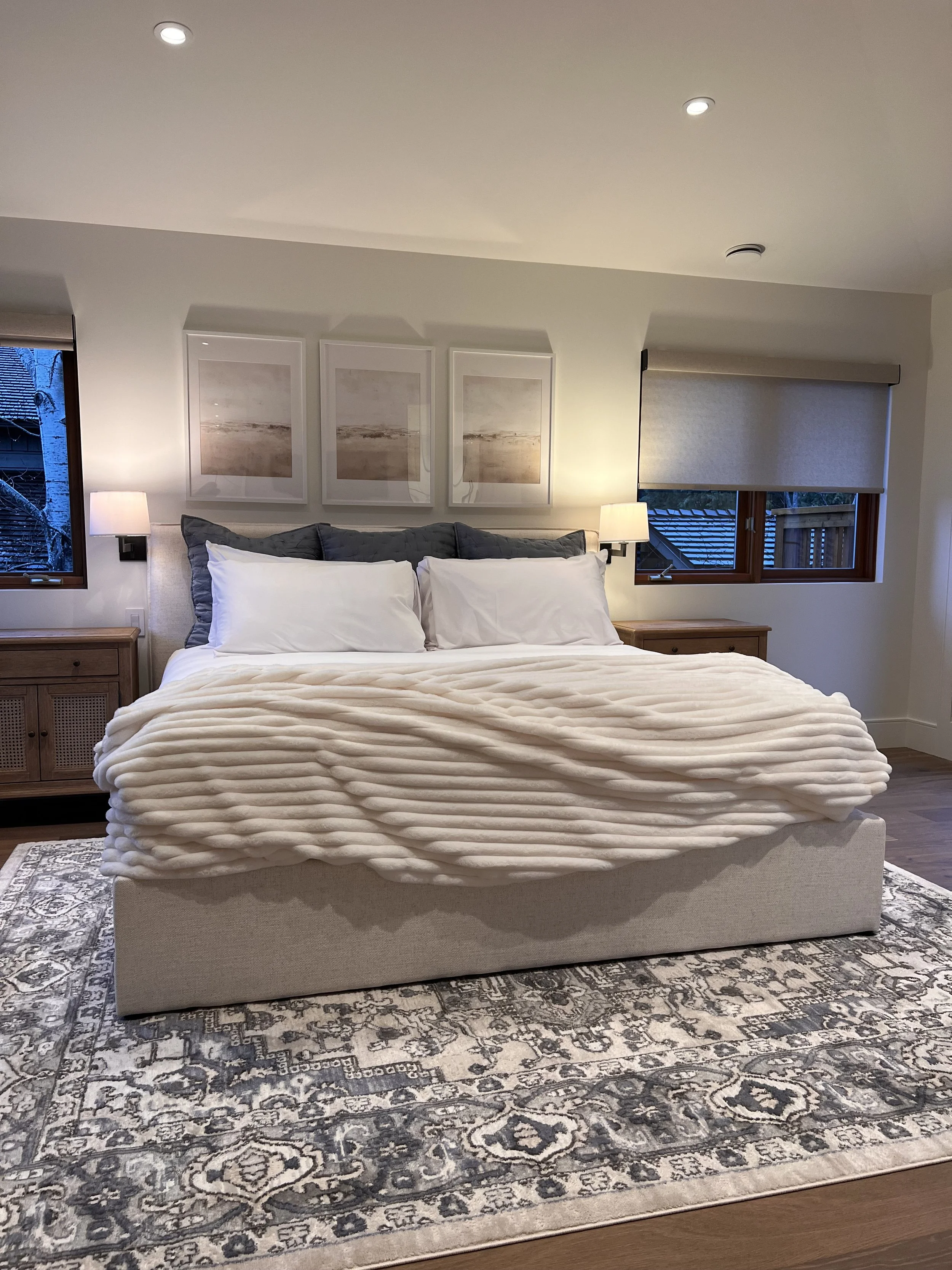 A neatly made bedroom with a king-sized bed, pillows, a textured cream blanket, and wooden nightstands. There are three framed pictures above the bed, two windows with blinds, and lamps on each side of the bed. The floor has a decorative rug.