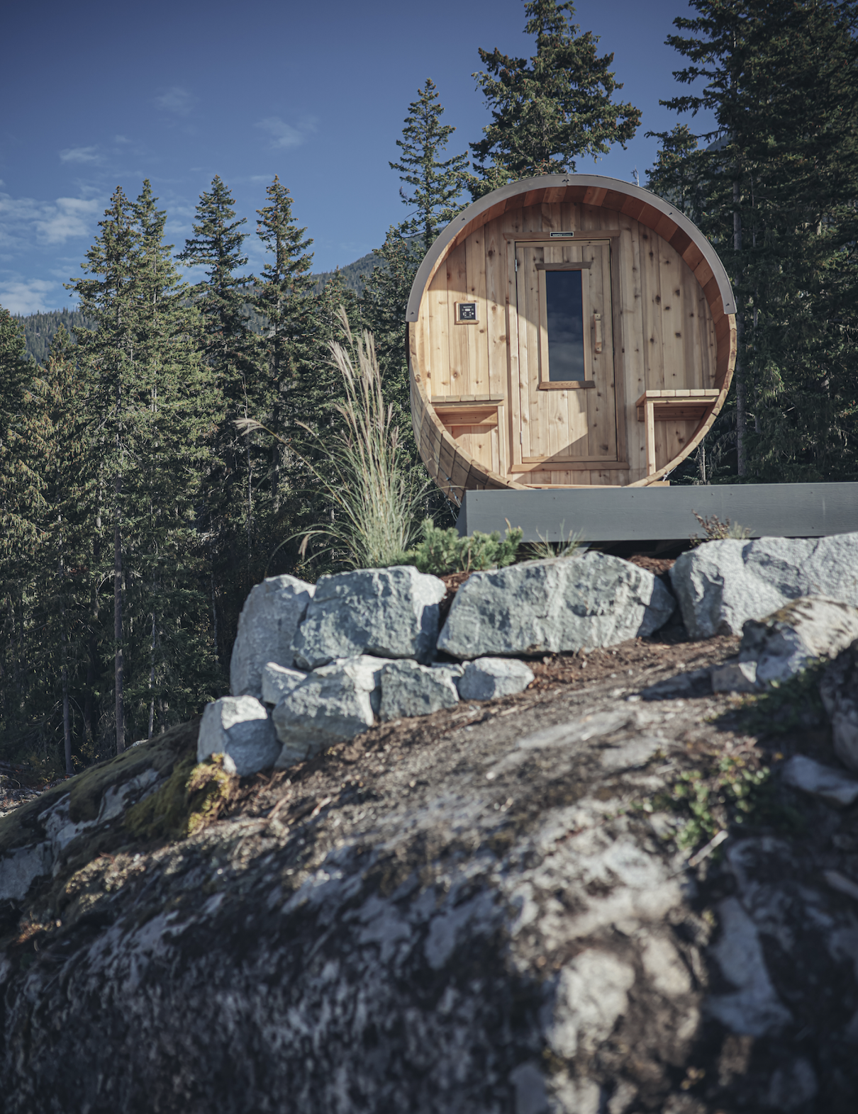 A small, round wooden sauna on a hilltop surrounded by trees with a rocky foreground and blue sky.