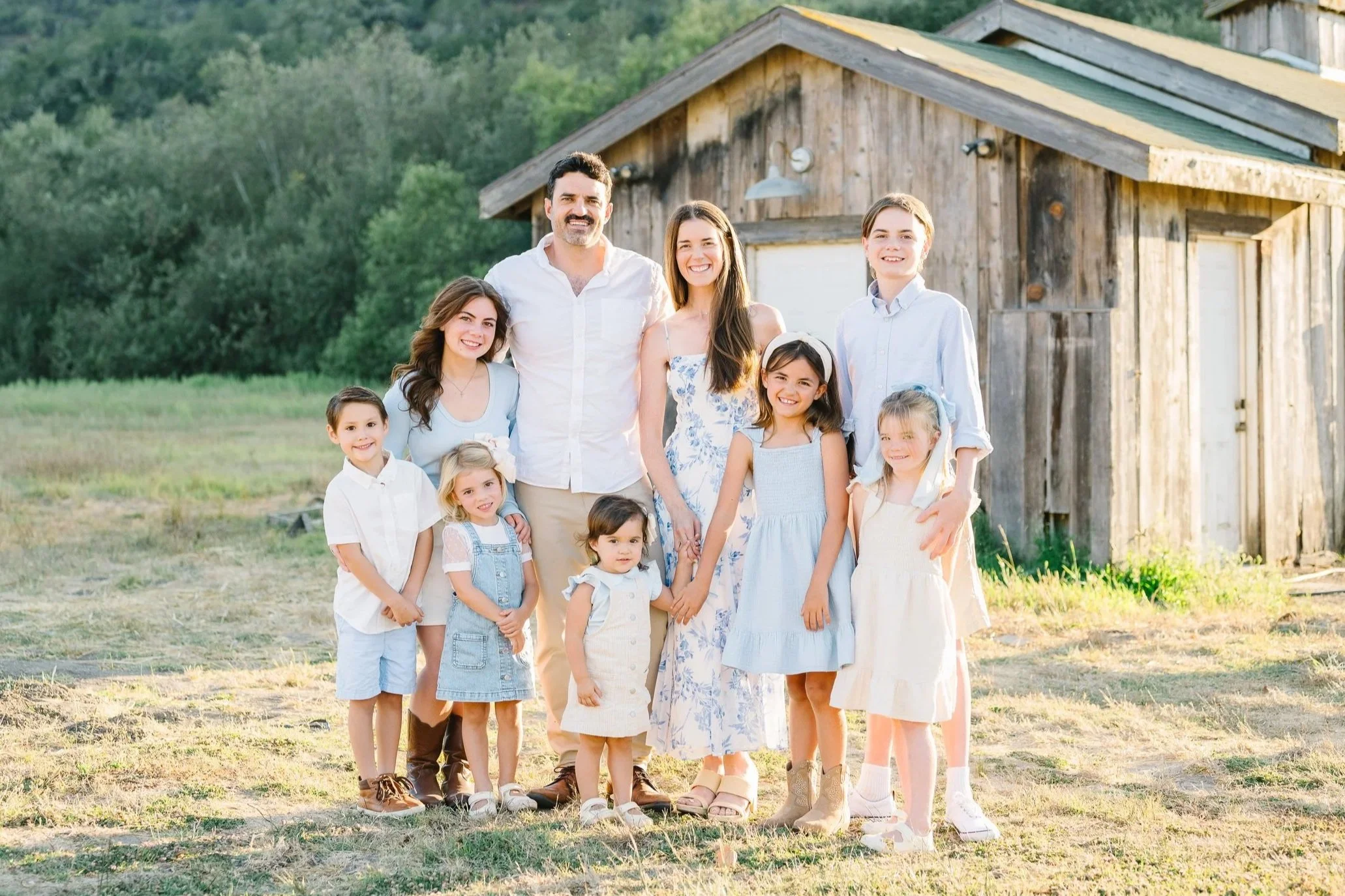 A family of ten standing outdoors in front of a wooden shed, smiling and dressed in light-colored clothing.