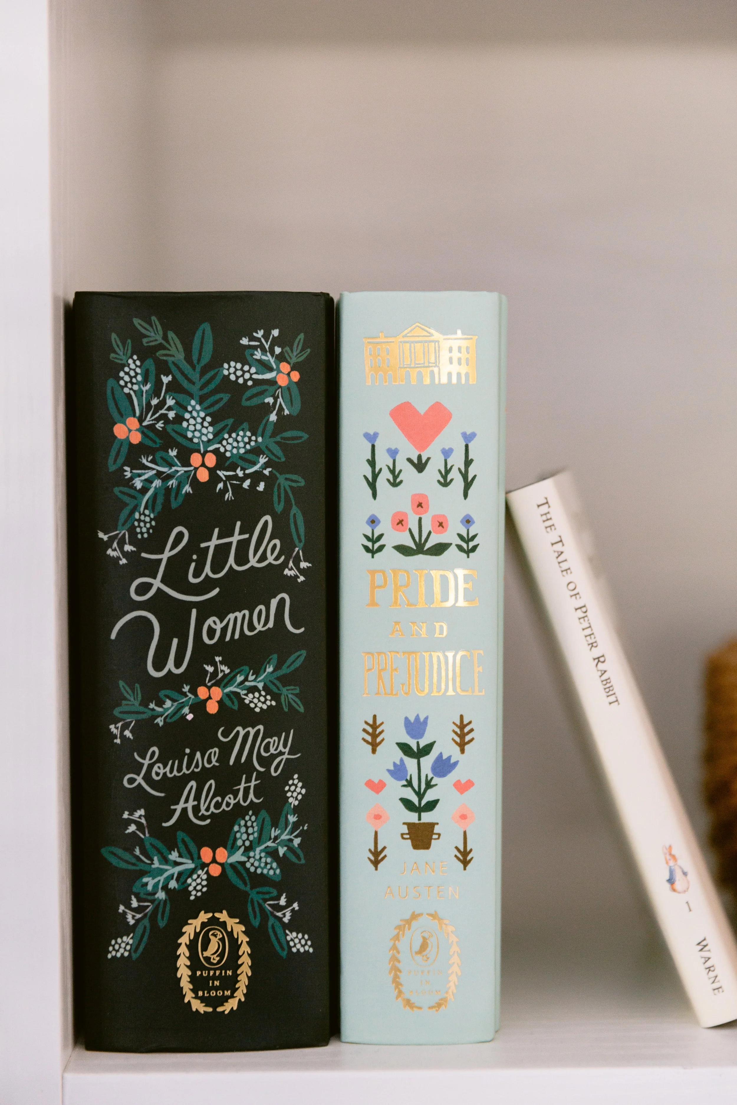 Two books on a shelf, one titled 'Little Women' by Louisa May Alcott and the other titled 'Pride and Prejudice' by Jane Austen, with a third smaller book titled 'The Tale of Peter Rabbit' visible nudging from the side.