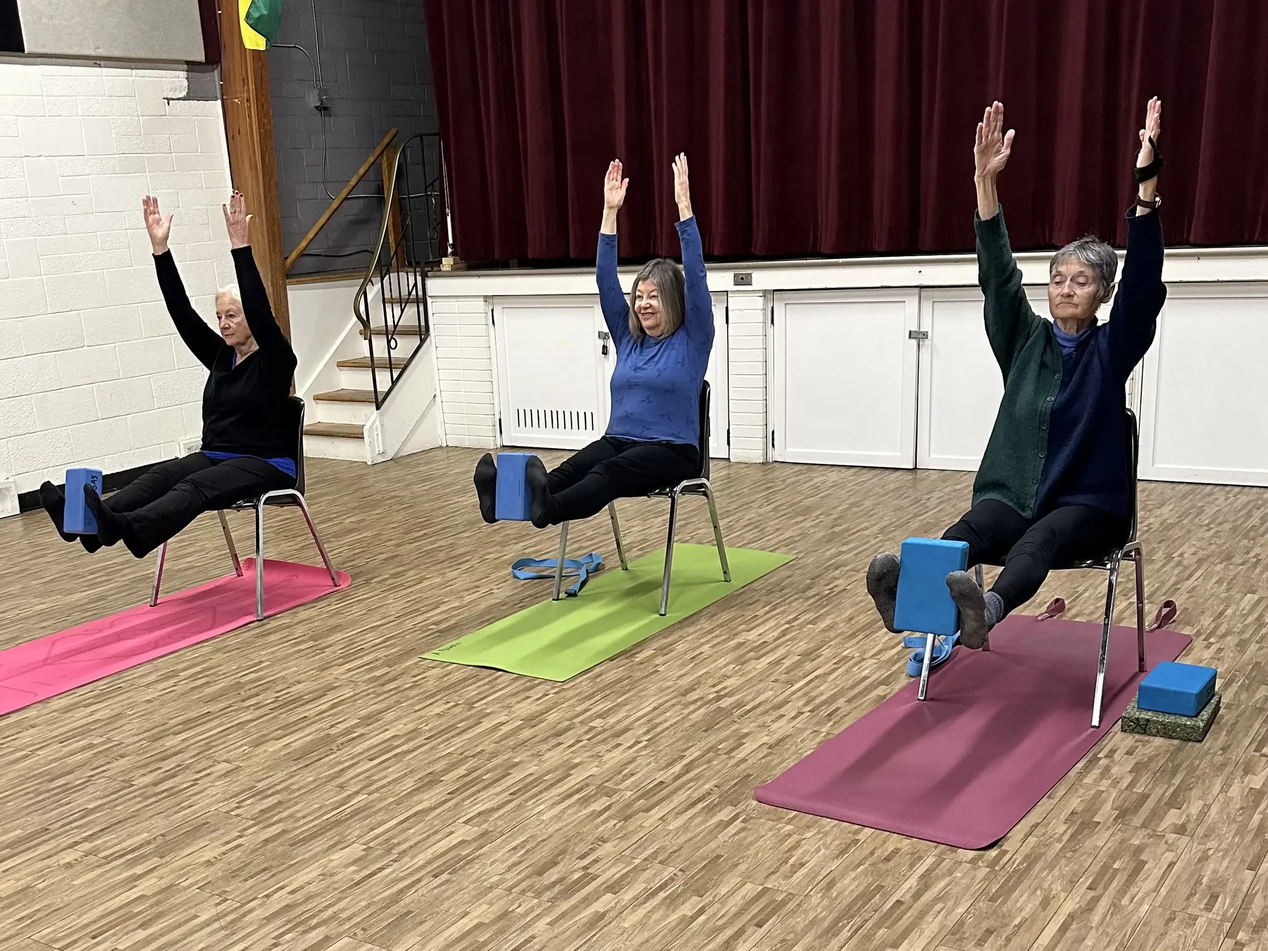 Three elderly women practicing chair yoga in a room with hardwood floors. They are seated in chairs with their arms raised above their heads, each on a different colored yoga mat with blue blocks on their laps.