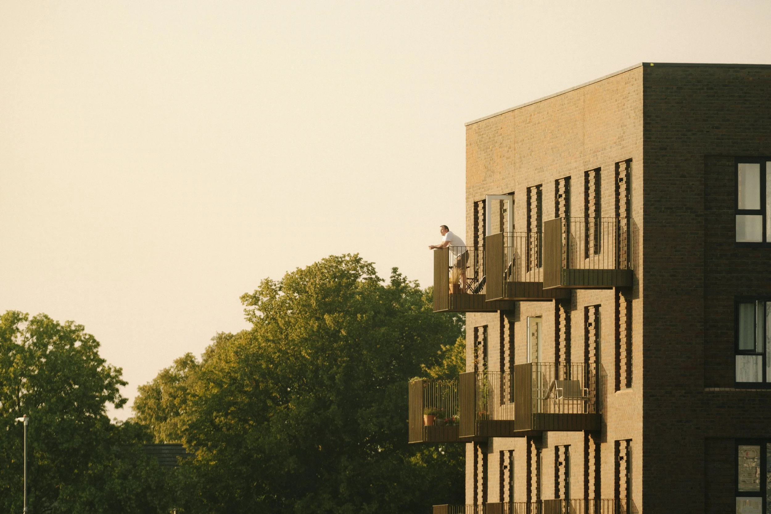A man sitting on a balcony of a modern brick apartment building, holding a cup, with trees and a clear sky in the background during sunset.