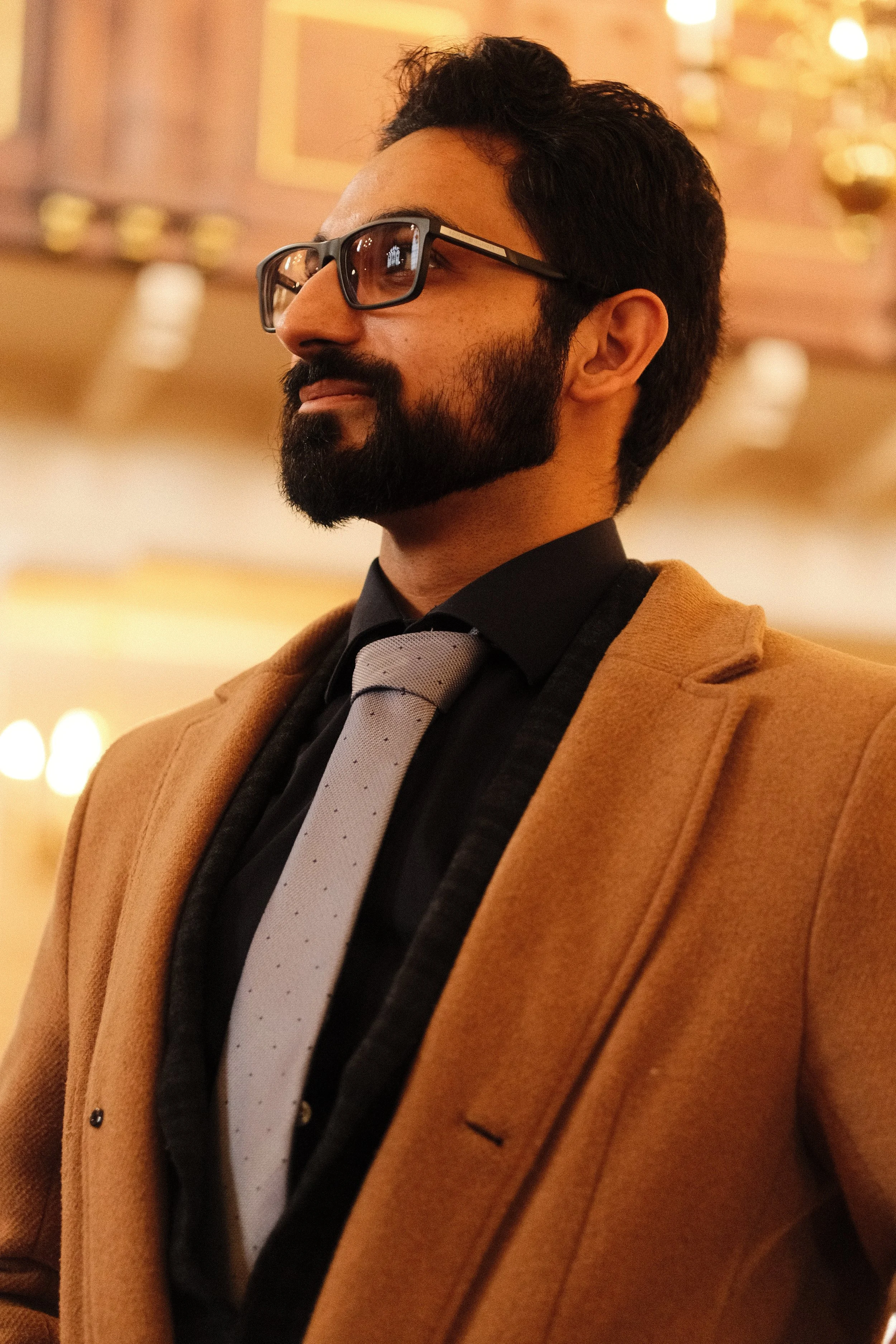 A man with a beard and glasses dressed in a tan coat, black shirt, and gray tie, standing indoors with warm lighting.