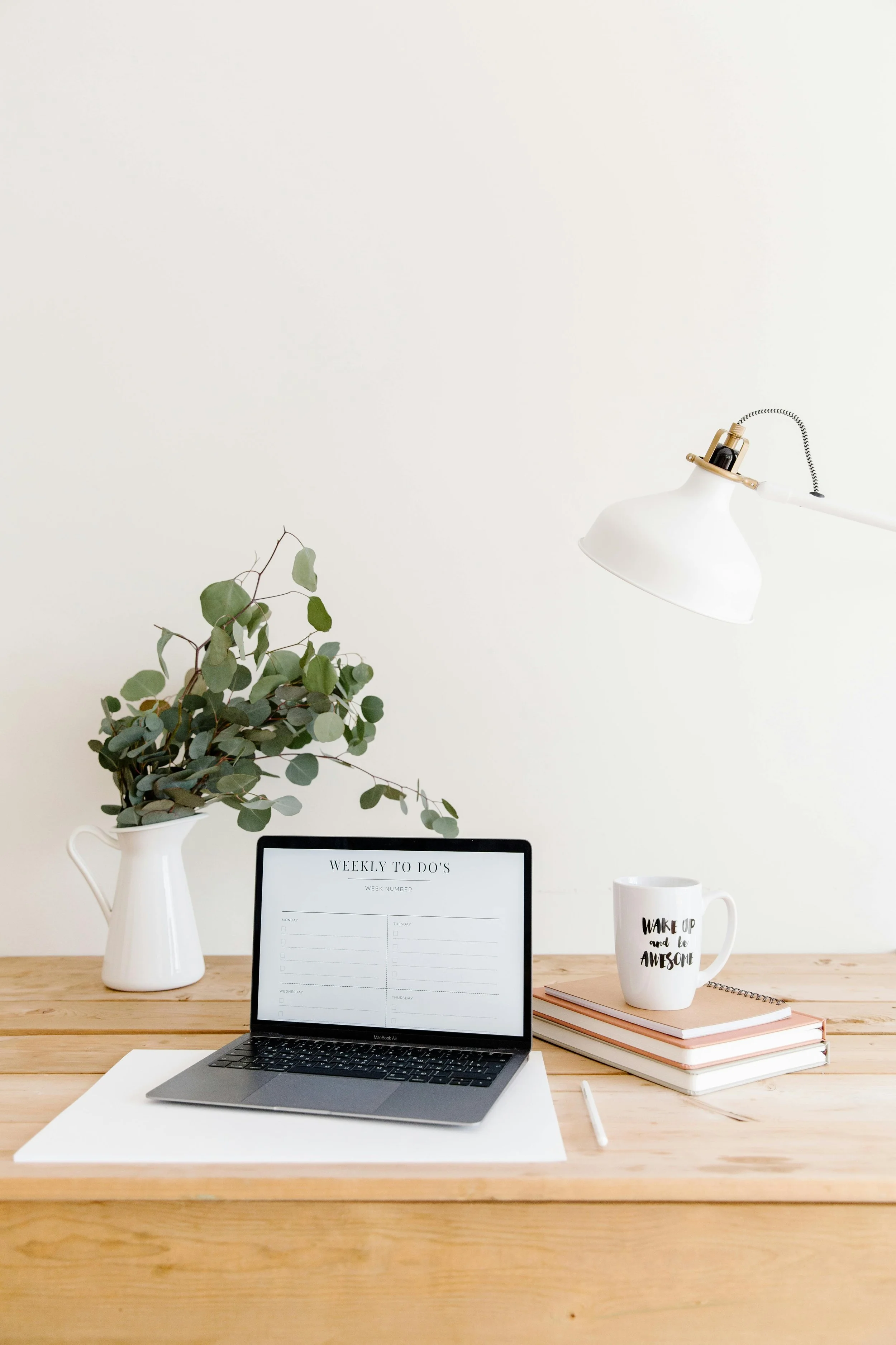 A tidy workspace with a wooden desk featuring a laptop, a white vase with greenery, a stack of notebooks, a mug with the phrase "Wake up and be awesome," a white paper on the desk, a pen, and a white desk lamp hanging above.