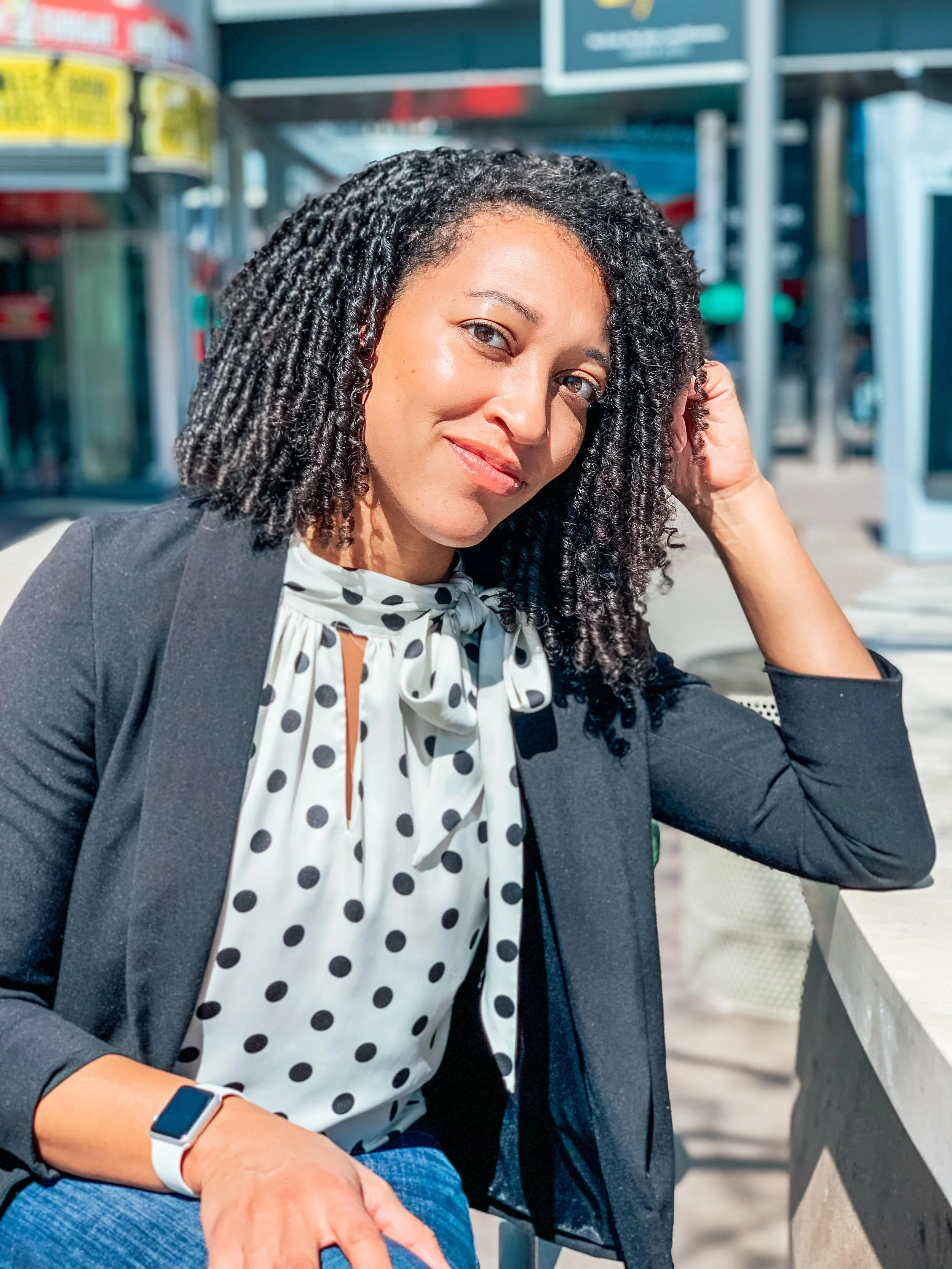 A woman with long, curly black hair sitting outdoors at a cafe table in bright sunlight, wearing a black blazer, white blouse with black polka dots and a bow, jeans, and a smartwatch.
