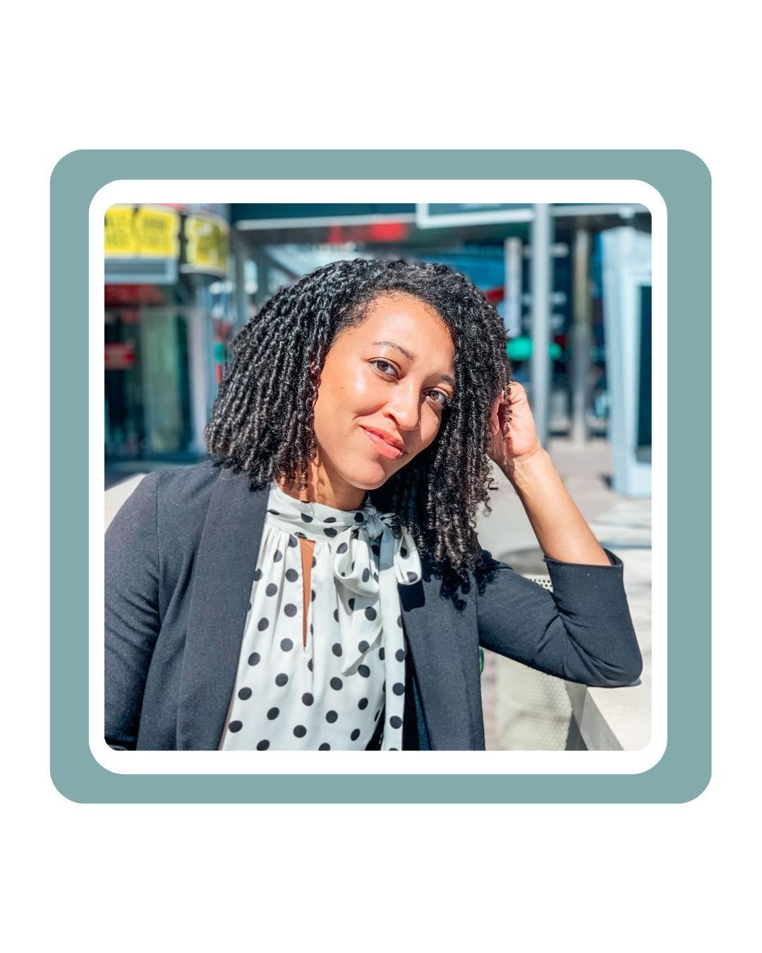 A woman with curly black hair wearing a polka dot blouse and blazer sitting outdoors.