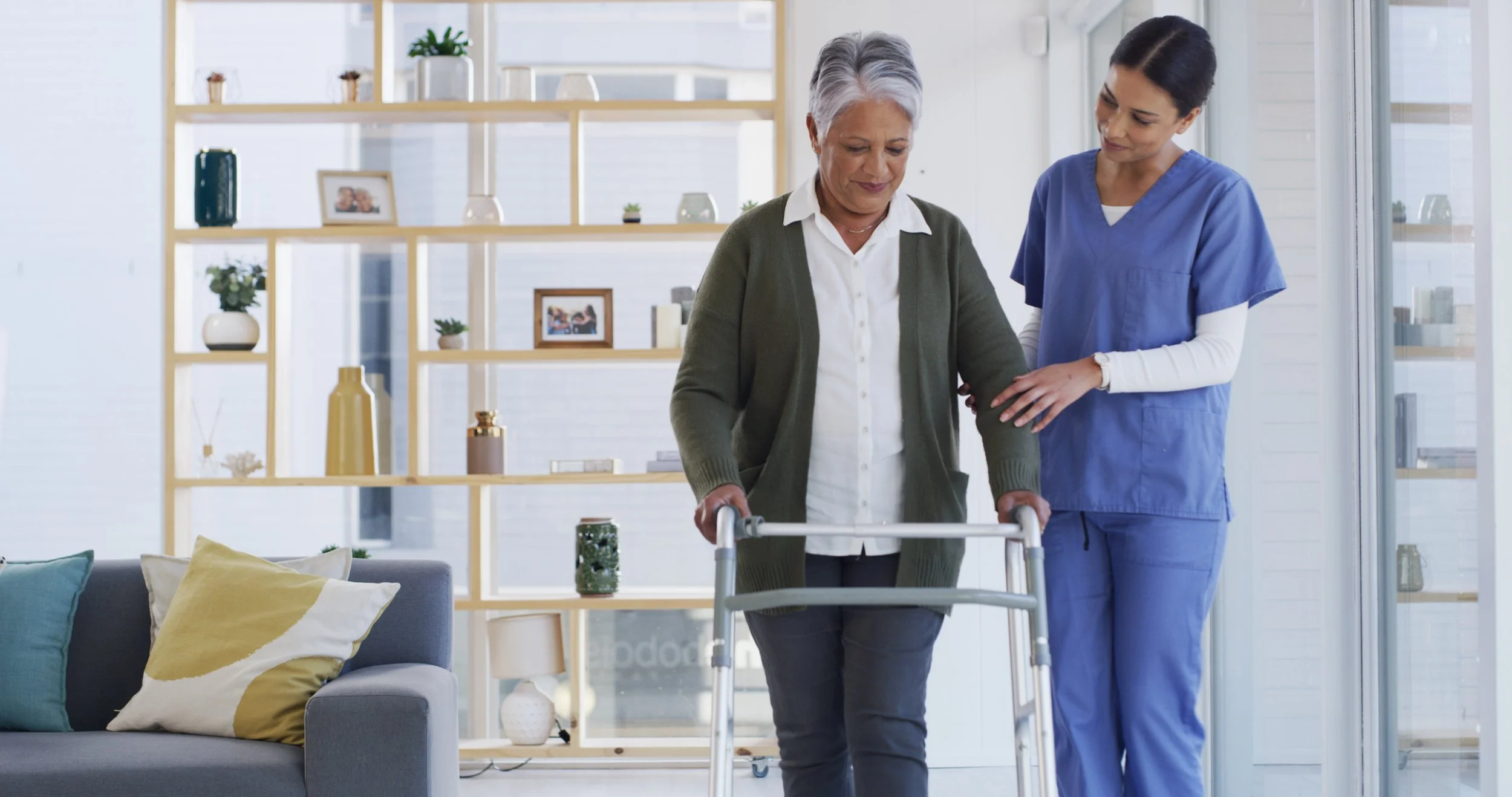An elderly woman with gray hair using a walker is assisted by a female healthcare worker in blue scrubs in a bright, modern living room.
